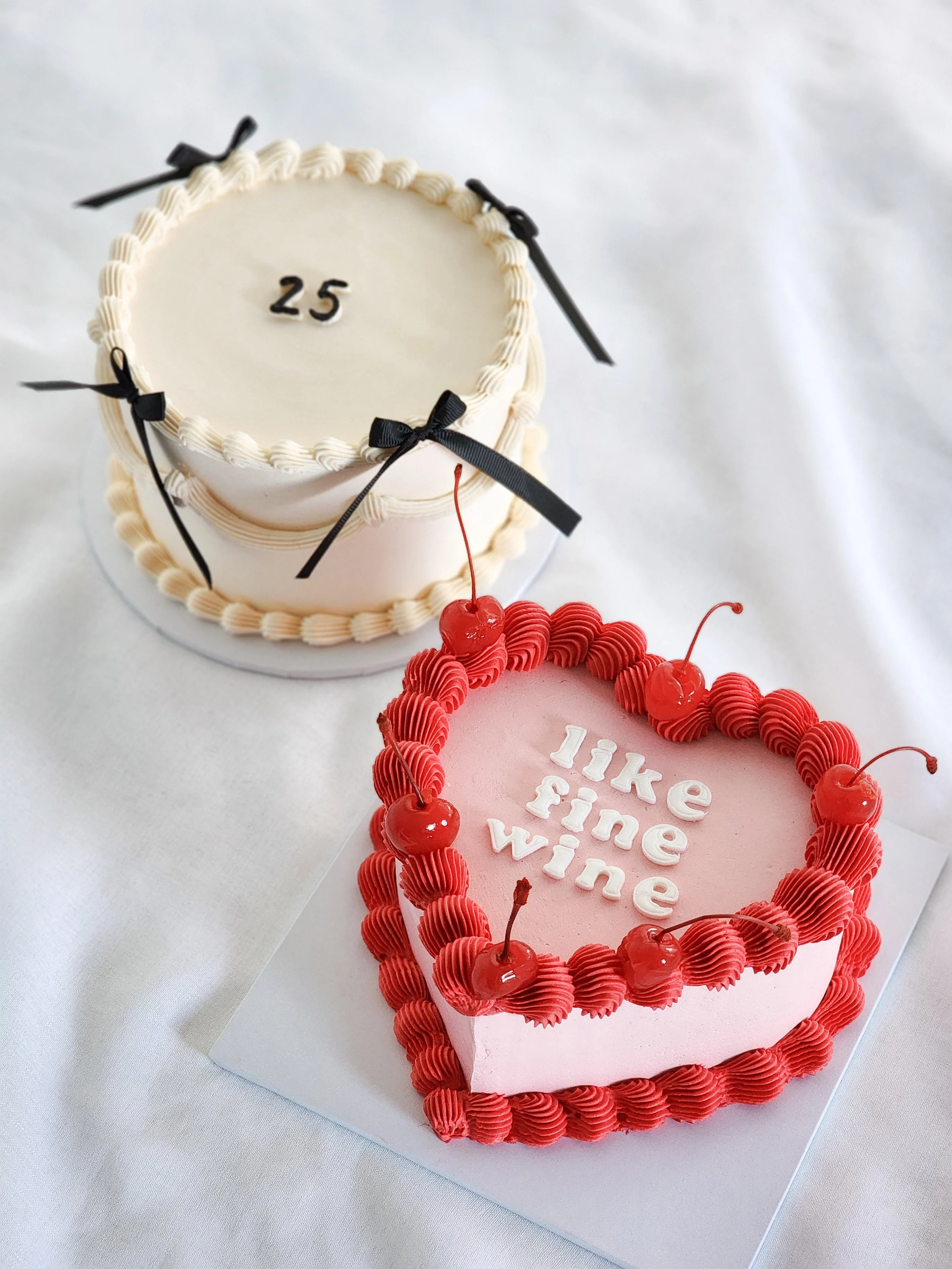 Two decorated birthday cakes, one white with black bows and the number 25 on top, and one pink with red frosting and cherries, with the text 'like fine wine'.