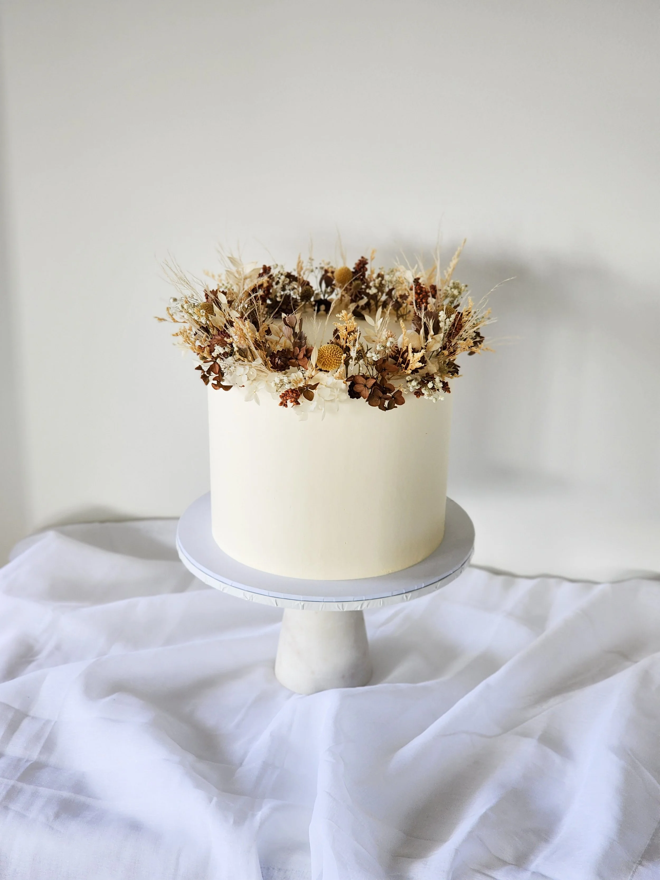 Cream-colored cake with dried floral arrangement on top, displayed on a white cake stand on a white draped fabric surface.