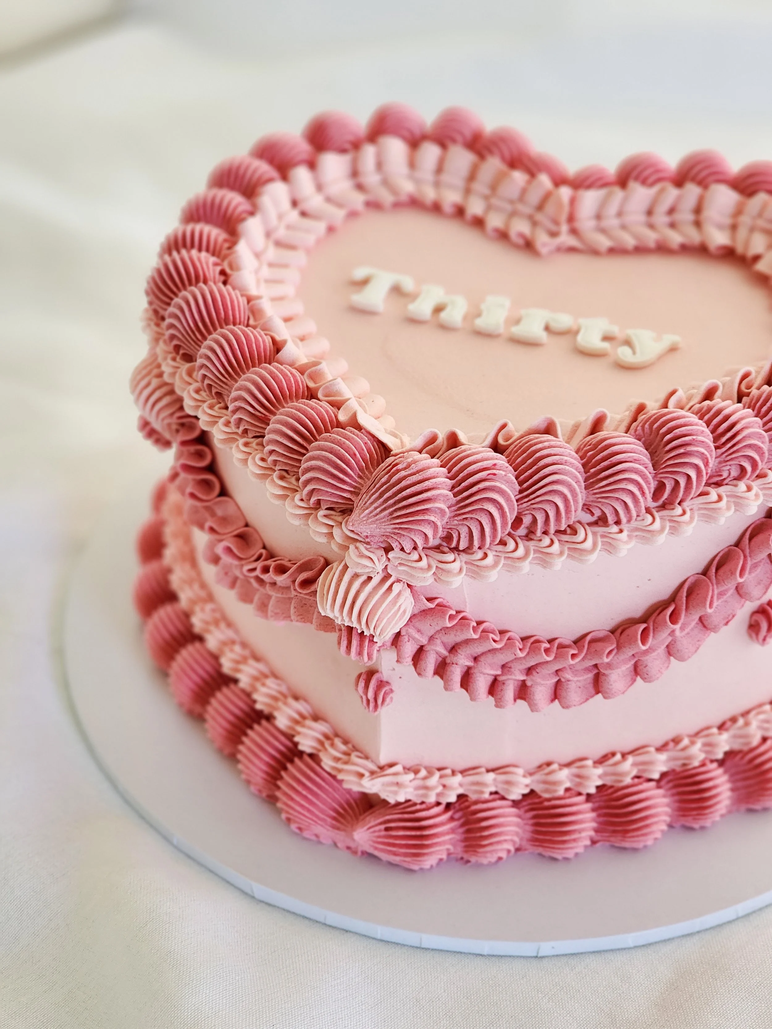 A pink heart-shaped cake decorated with pink frosting and piped swirls, with the words "Thirty" written in white icing on top.