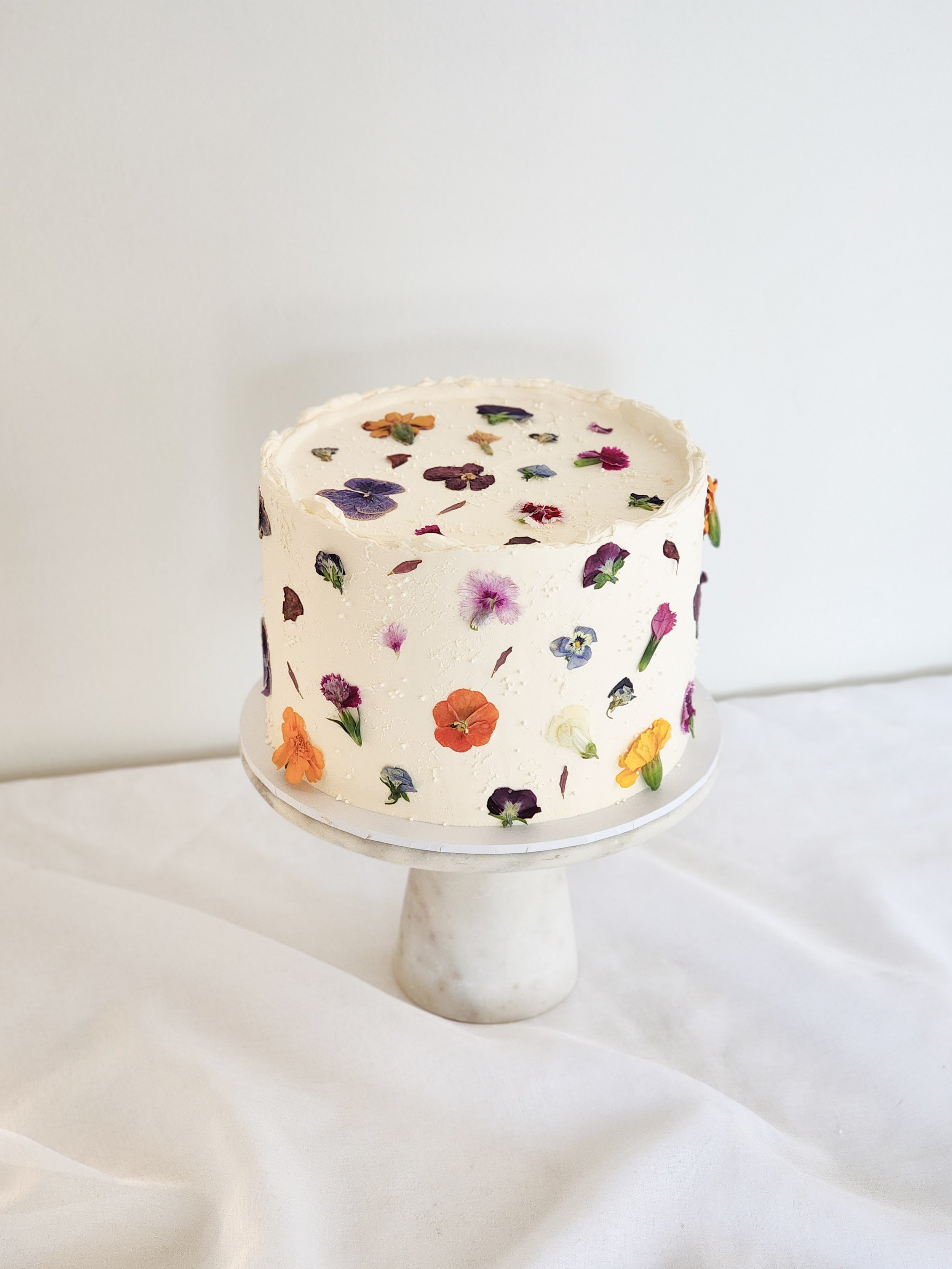 A white frosted cake decorated with various colorful edible flowers, on a marble cake stand against a white background.