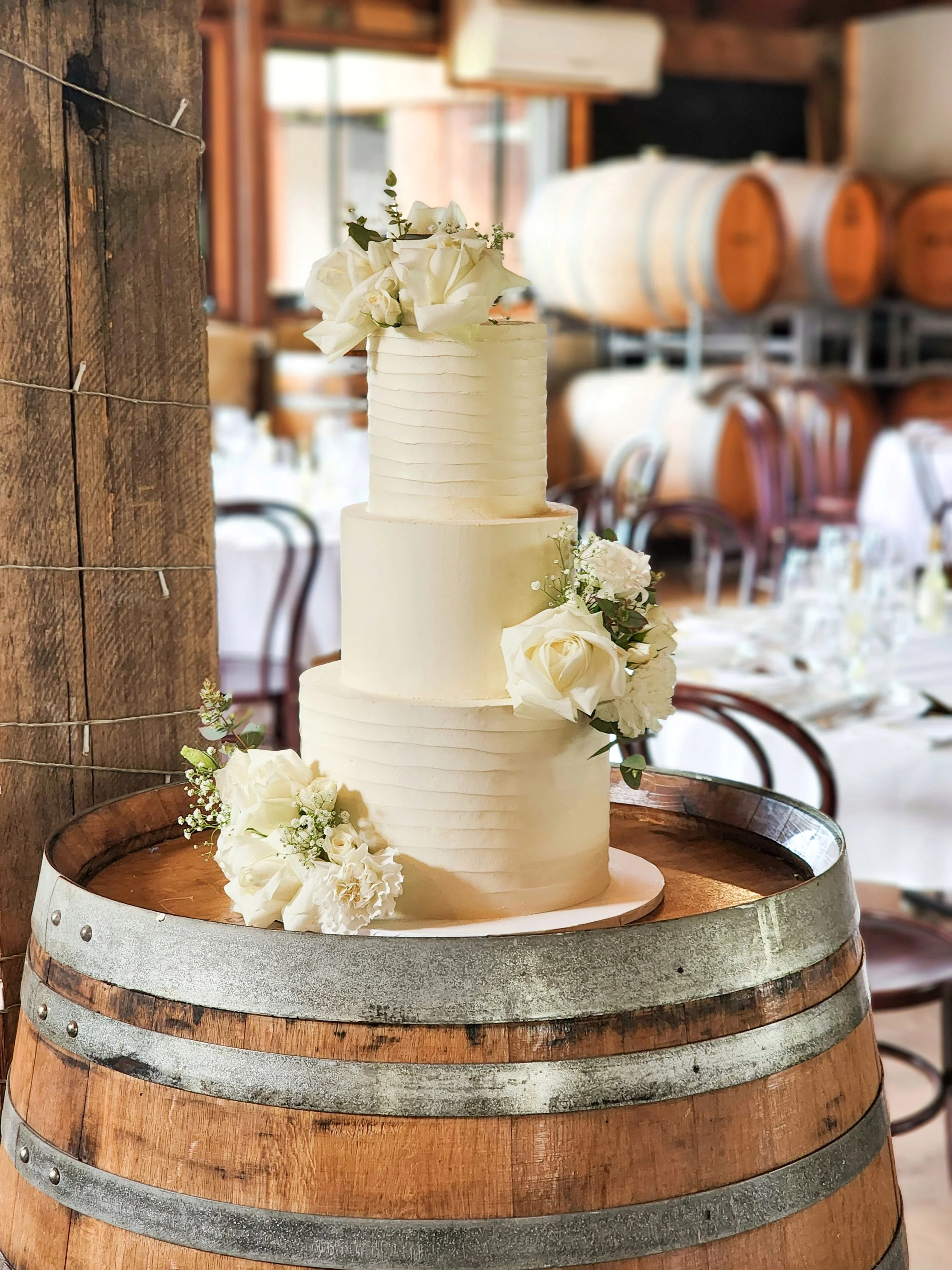 Three-tier white wedding cake with floral decorations on a rustic wooden barrel in a winery setting.