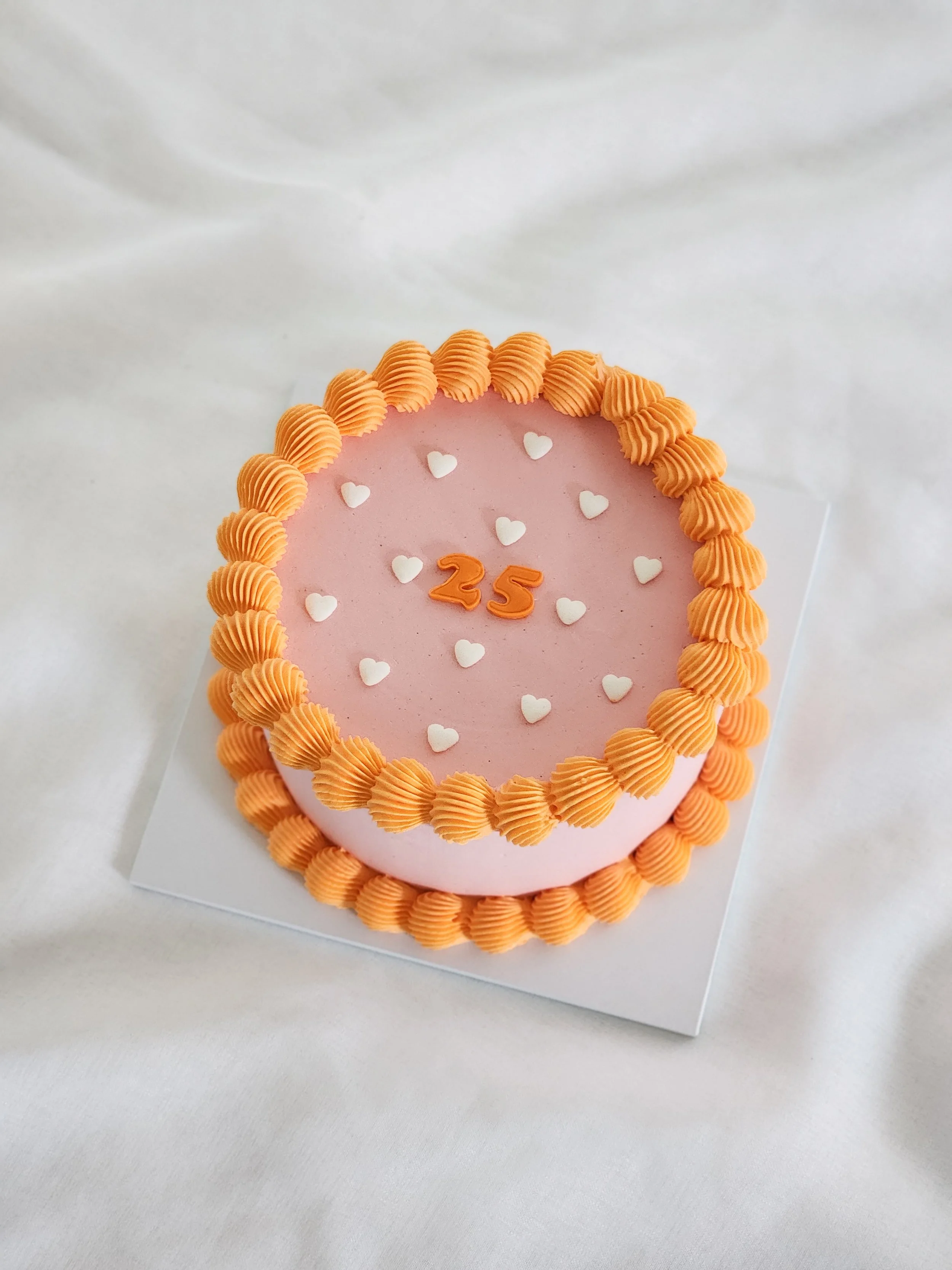 Pink birthday cake with orange border and small white hearts, decorated for a 25th birthday celebration.