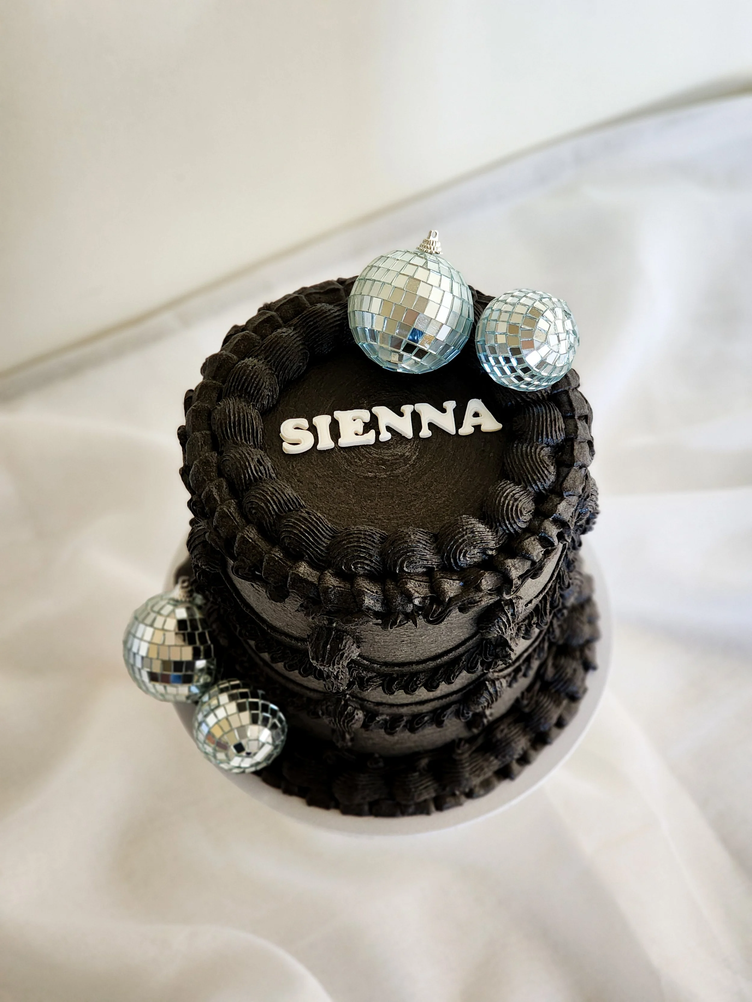 A small chocolate cake with black frosting and decorative piping, topped with white letters spelling 'SIENNA' and three disco ball ornaments, on a white cake board.