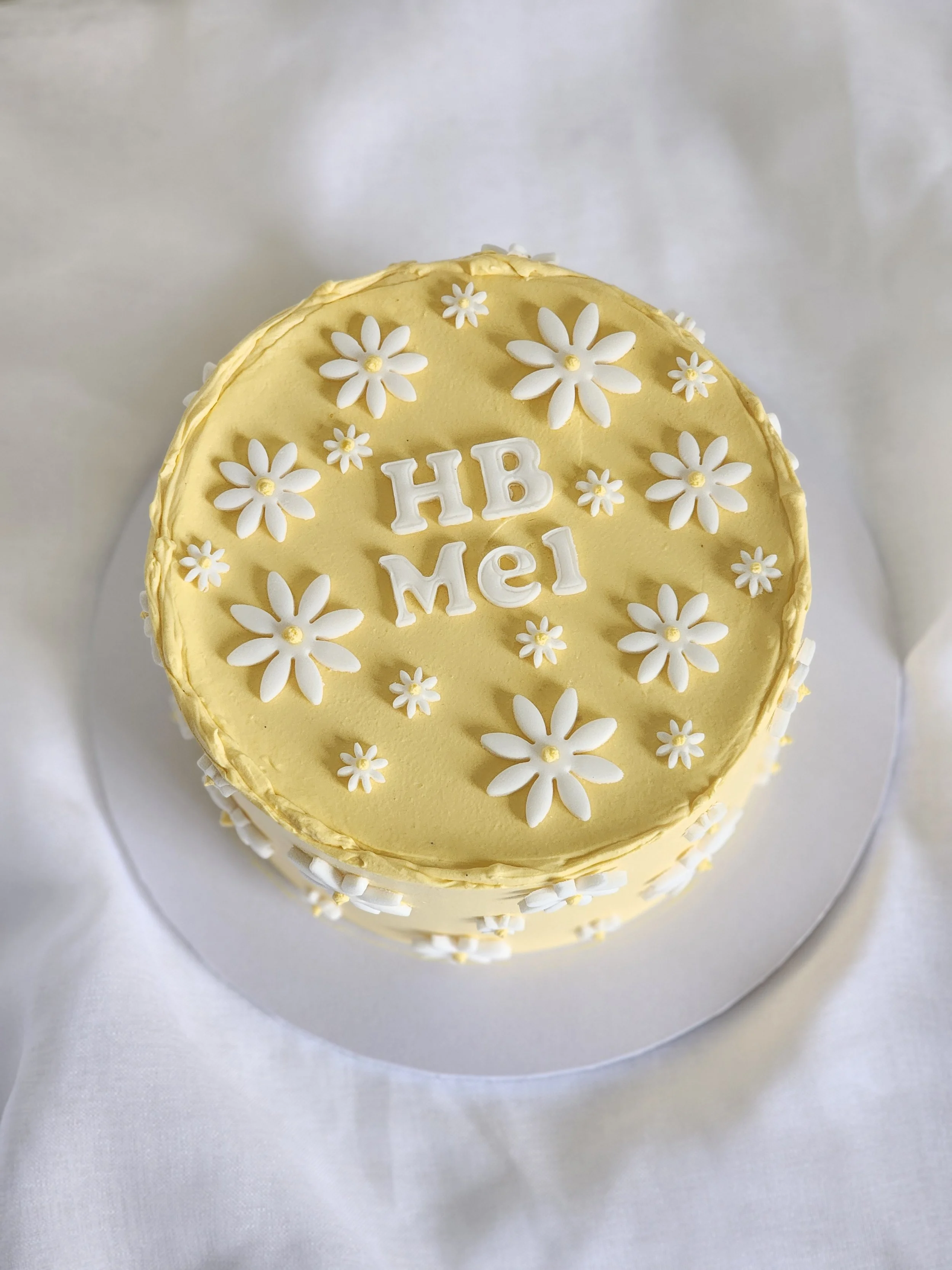 Yellow birthday cake decorated with white flowers and the words "HB Mel" on top.