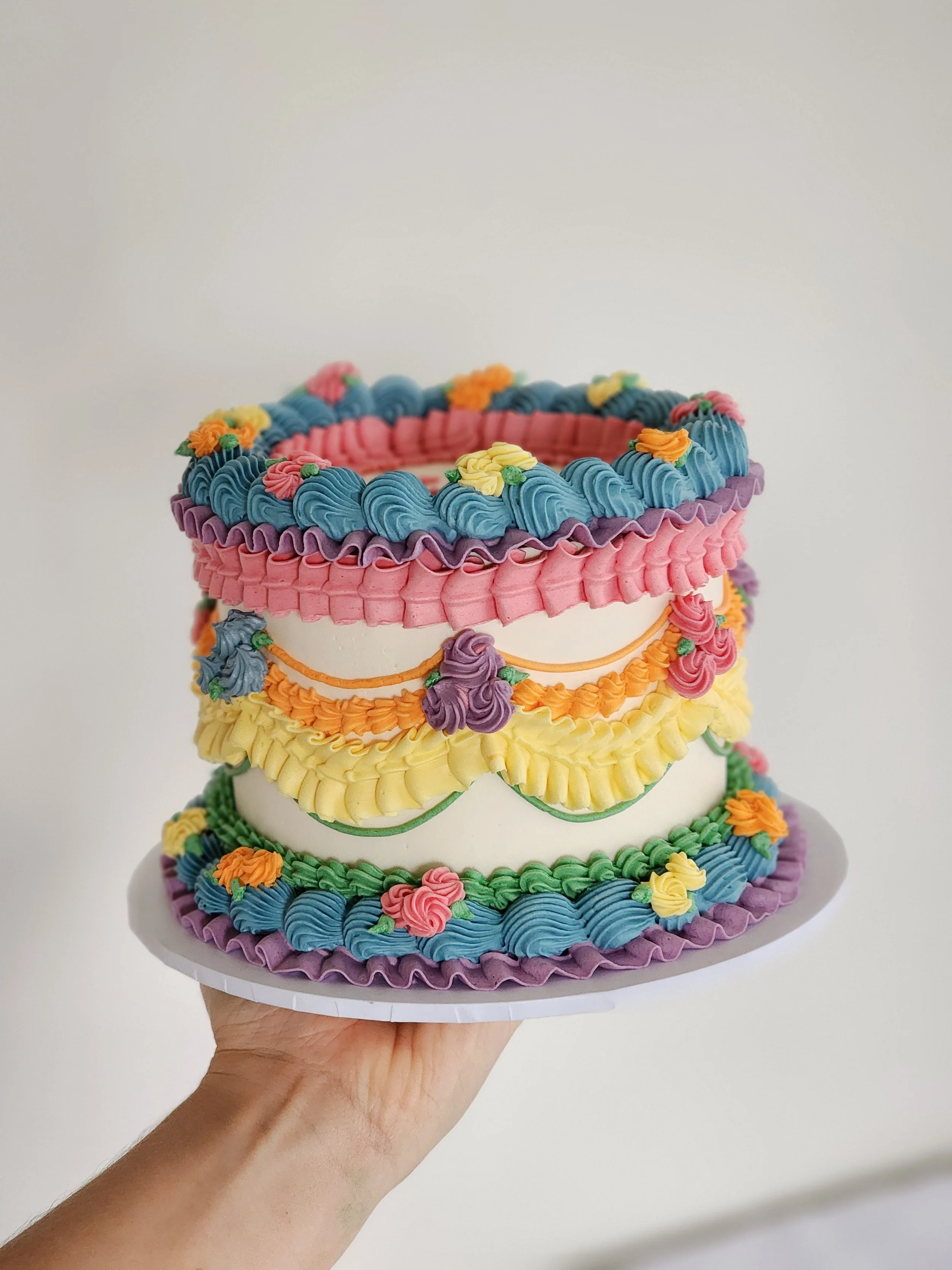 Colorful rainbow vintage birthday cake decorated with pink, yellow, green, purple, blue, and orange icing flowers and swirls, held on a white cake board.