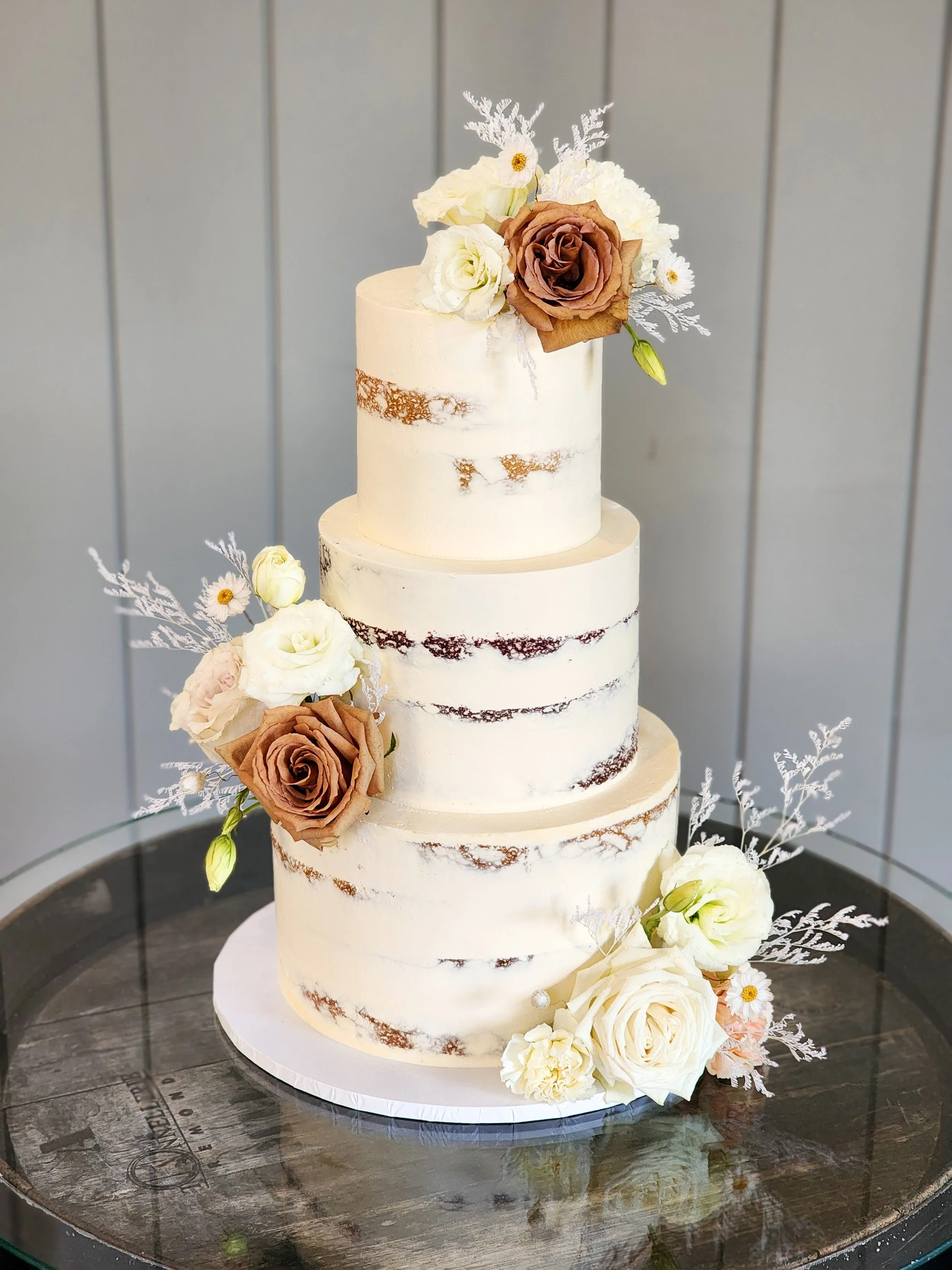 Three-tier naked cake with white frosting and decorated with white and brown roses on a round glass table.