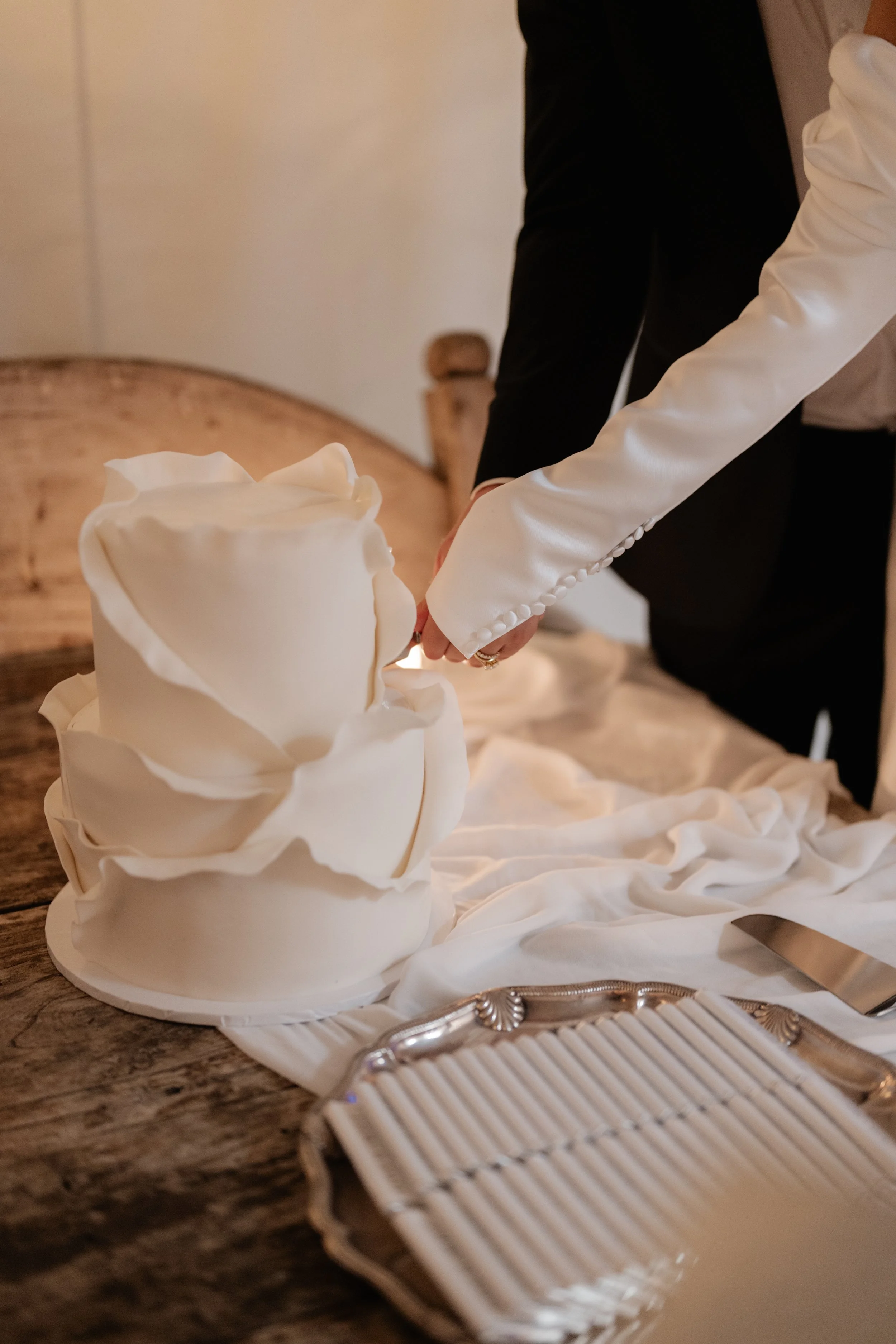A white tiered cake with decorative ruffles, a hand in a white satin glove holding a knife, a silver tray, and a white cloth on a wooden table.