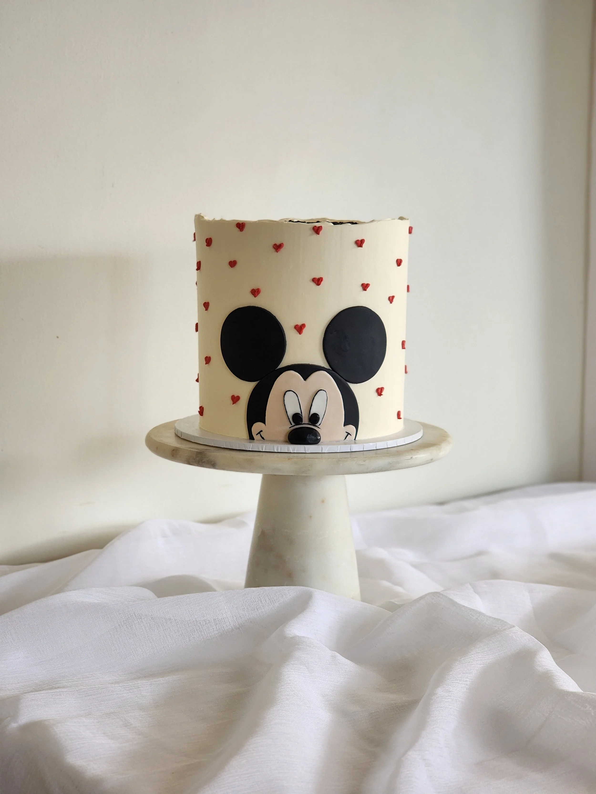 A Mickey Mouse themed cake with small red hearts, placed on a white cake stand, with a white cloth in the foreground.