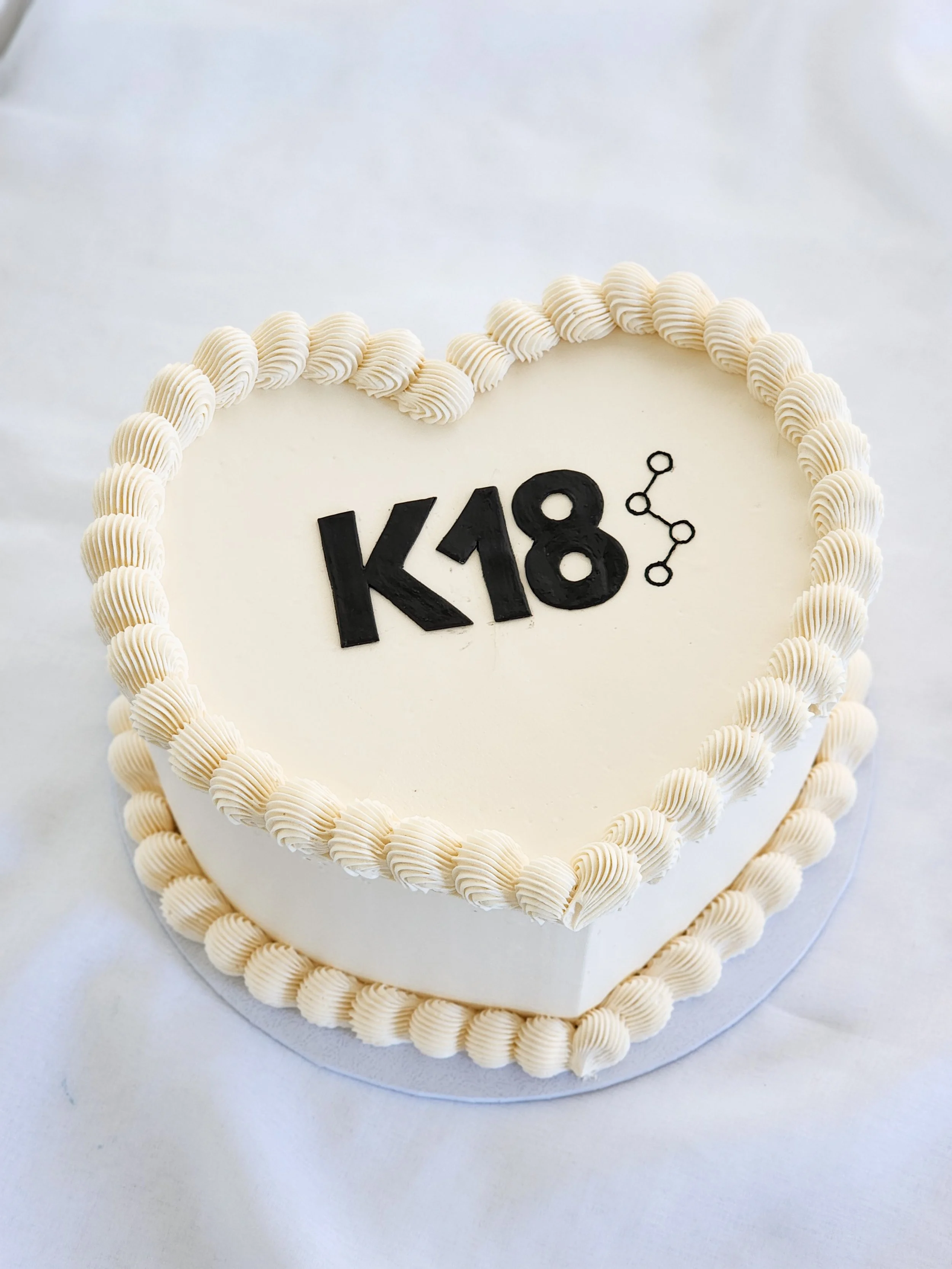 Heart-shaped cake with white icing, decorated with white piped border, featuring the text 'K18' and a small constellation symbol.