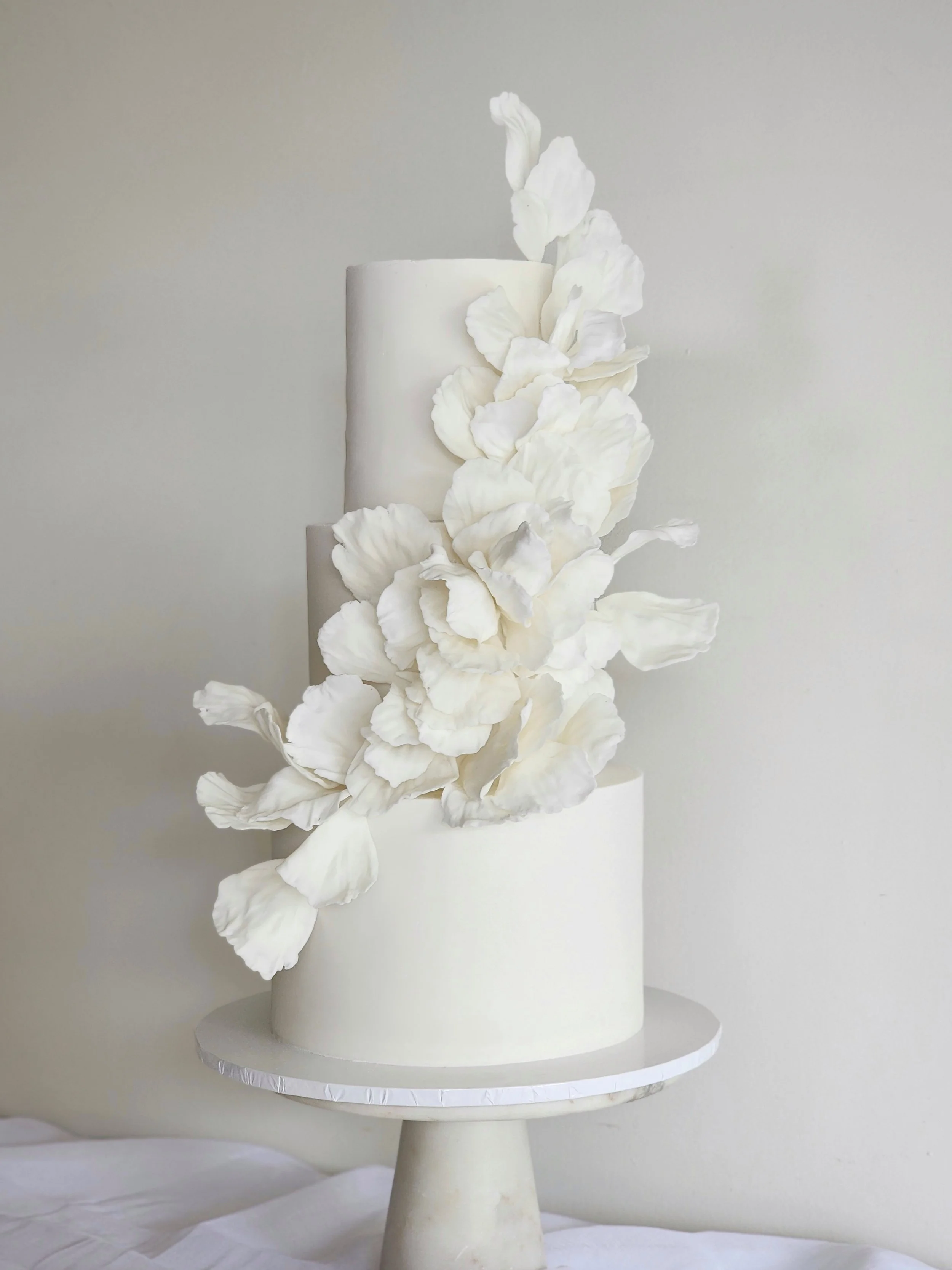 Elegant three-tier white wedding cake with delicate white floral decorations cascading down the side, displayed on a round pedestal stand.