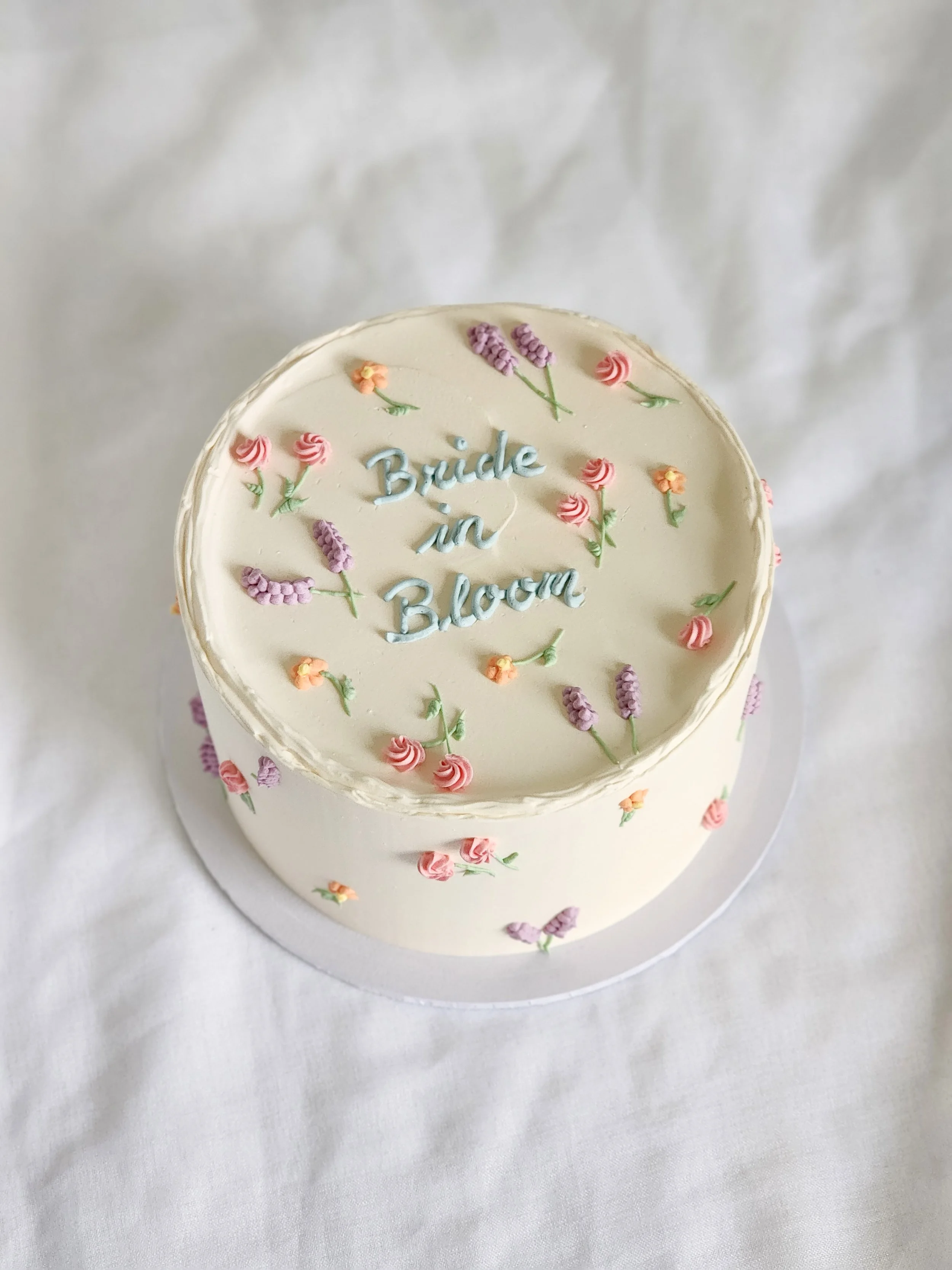 A round bridal shower cake with white frosting decorated with small pink, purple, and orange flowers. The top of the cake has the words "Bride in Bloom" written in blue icing, surrounded by additional small flowers.