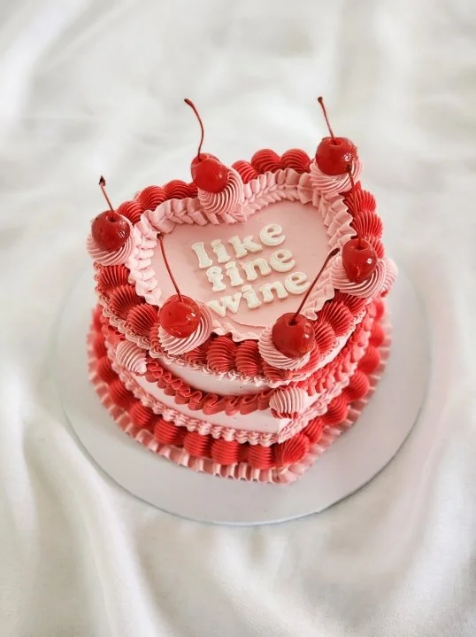 Heart-shaped pink and red layered cake with cherries on top and the message "like fine wine" in the center.