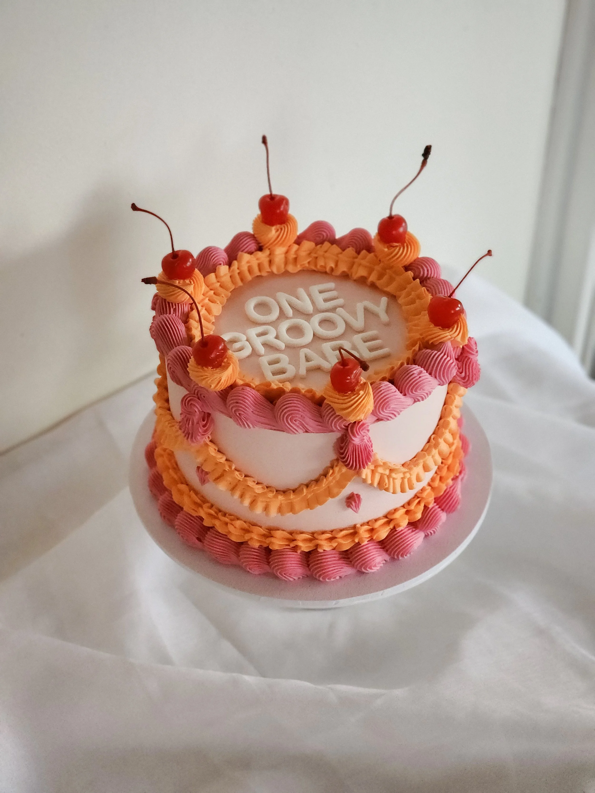 A birthday cake decorated with pink and orange frosting, topped with cherries, with the message "One Groovy Baby" written on top.
