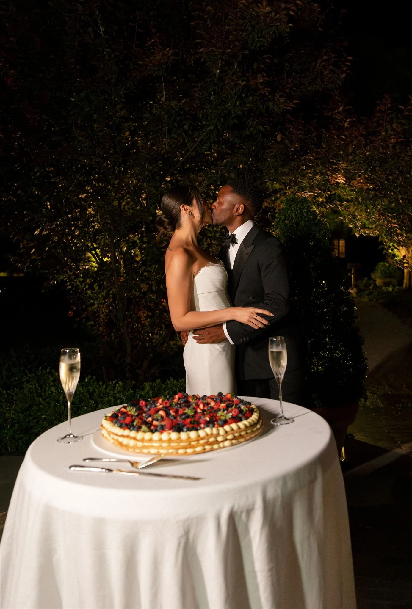 A newlywed couple kissing at their wedding reception, decorated outdoors at night with trees and warm lighting. In front of them is a table with a large fruit-topped wedding Millefoglie / Millefeuille cake, two champagne glasses, and silverware.
