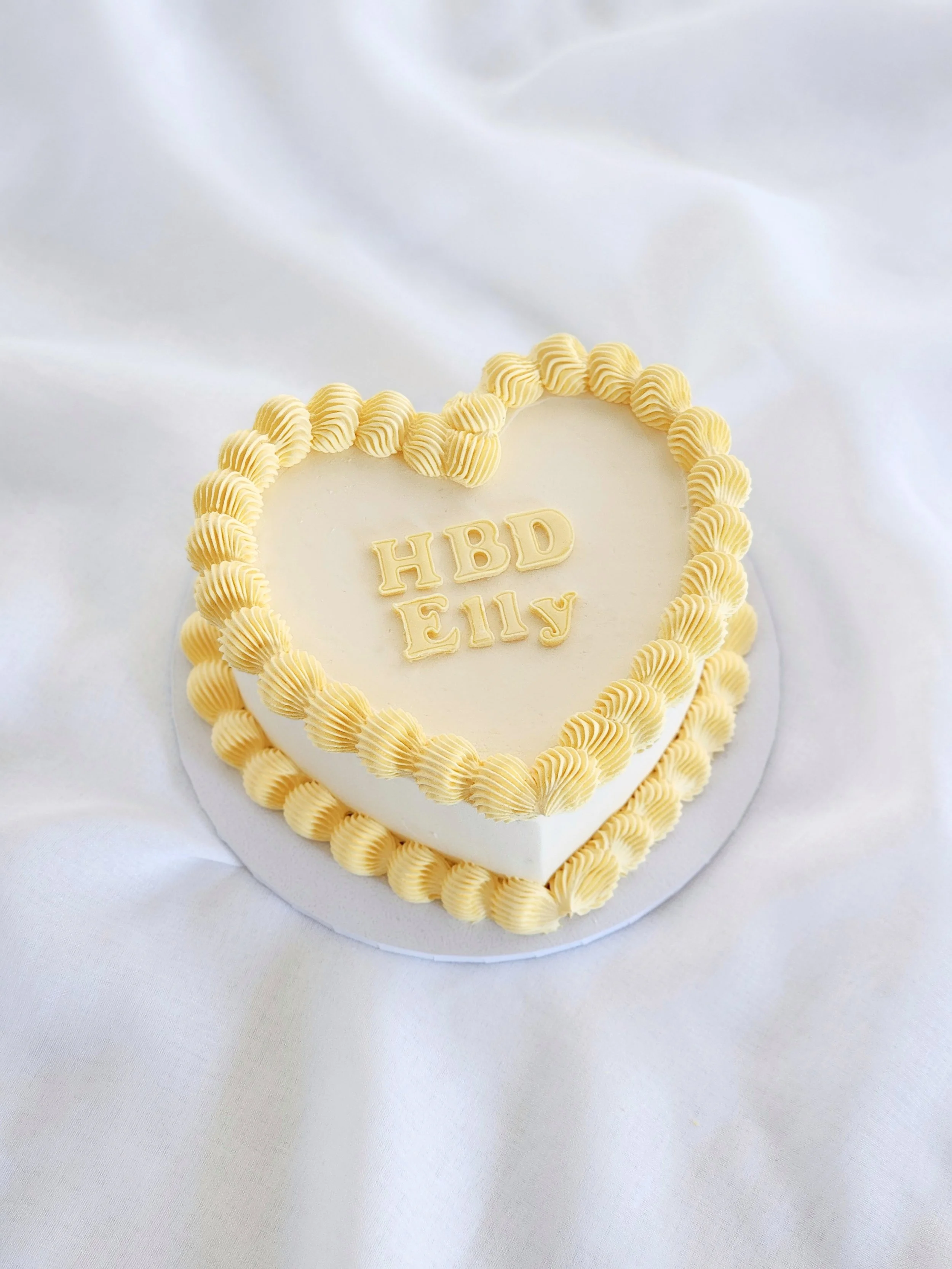 Heart-shaped birthday cake with yellow frosting border and written text 'HBD Elly' on top, placed on a white surface.