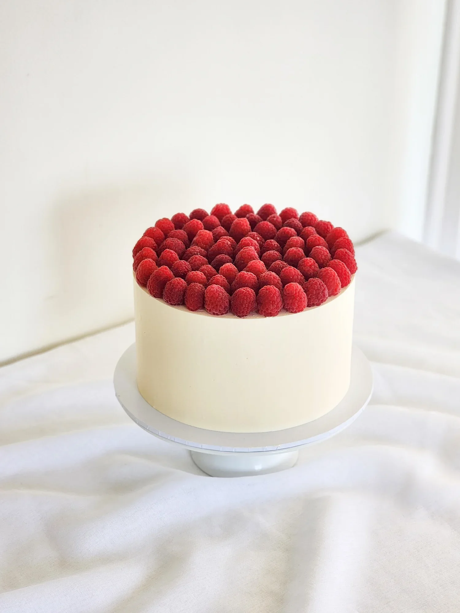 White cake topped with a border of fresh raspberries on a white cake stand, against a white background.