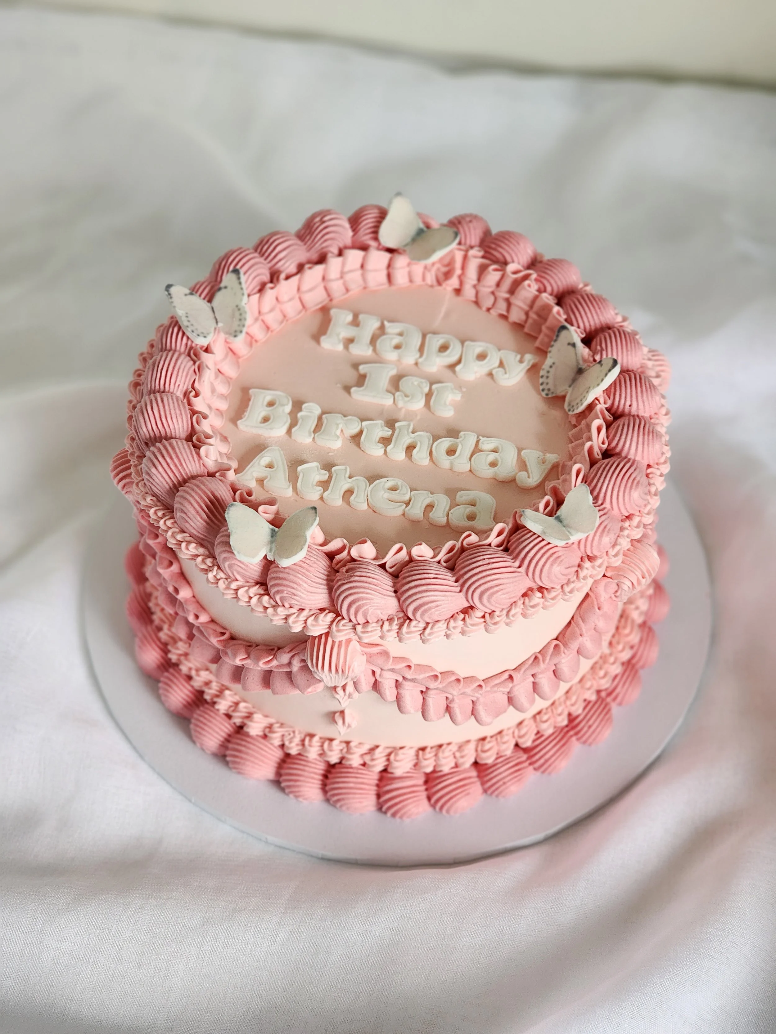 Pink birthday cake decorated with white butterflies and pink rosettes, with the message 'Happy 1st Birthday Athena' written on top.