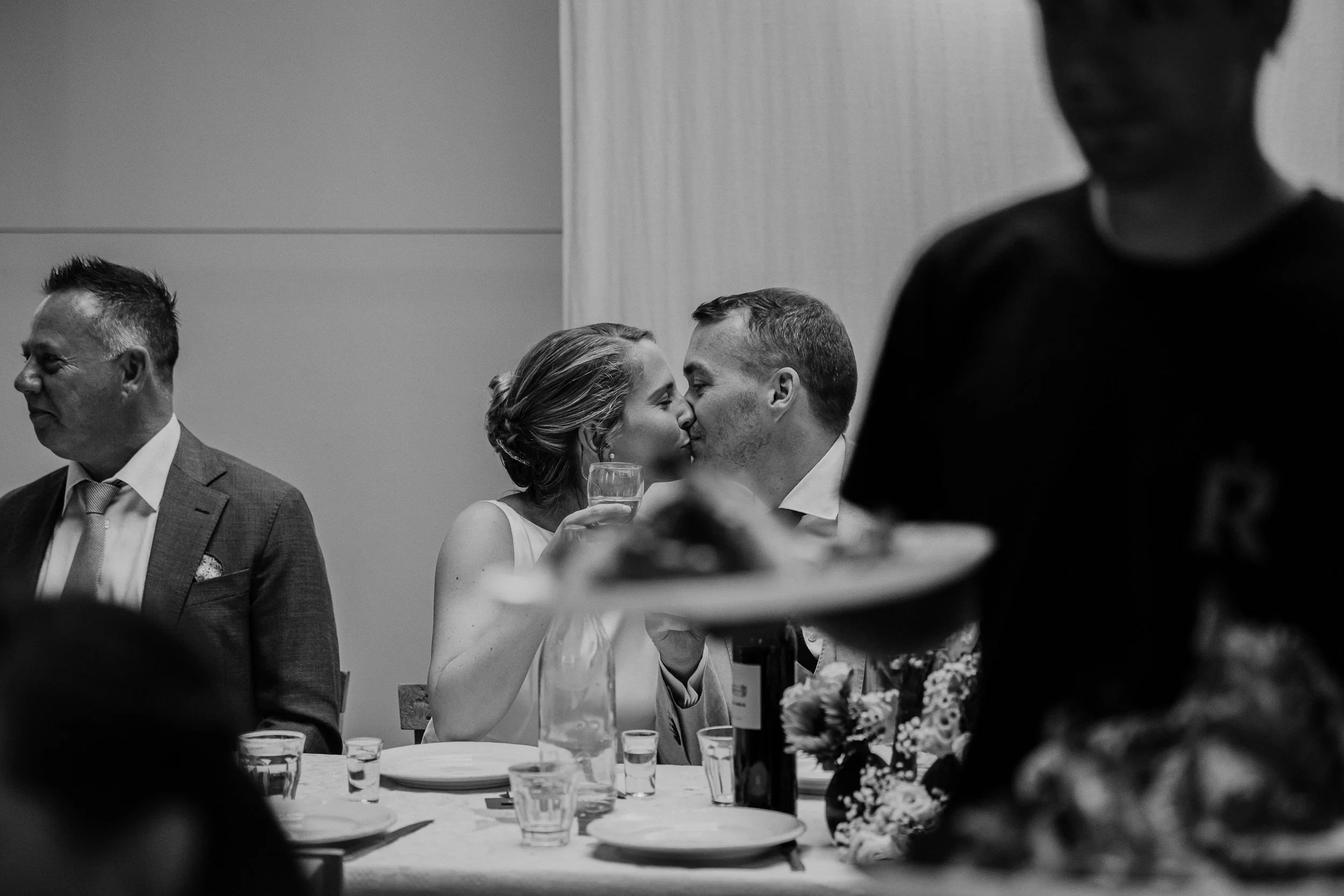 Bruidspaar kust elkaar tijdens het diner in trouwlocatie het Rijk van de Keizer in Amsterdam. Spontane zwart wit foto.