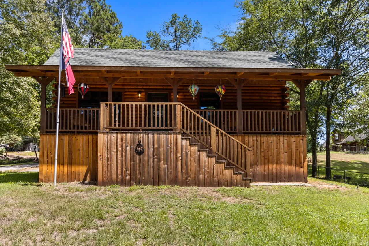 A wooden house with a raised porch, stairs, trees in the background, and an American flag on a flagpole in the front yard.