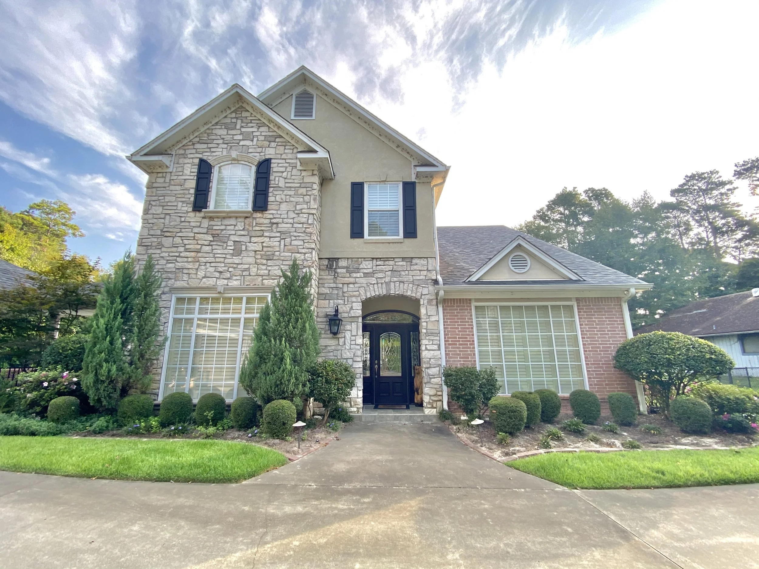 Front view of a two-story house with a stone and brick exterior, large windows, and a black front door, surrounded by a manicured garden and driveway under a partly cloudy sky.