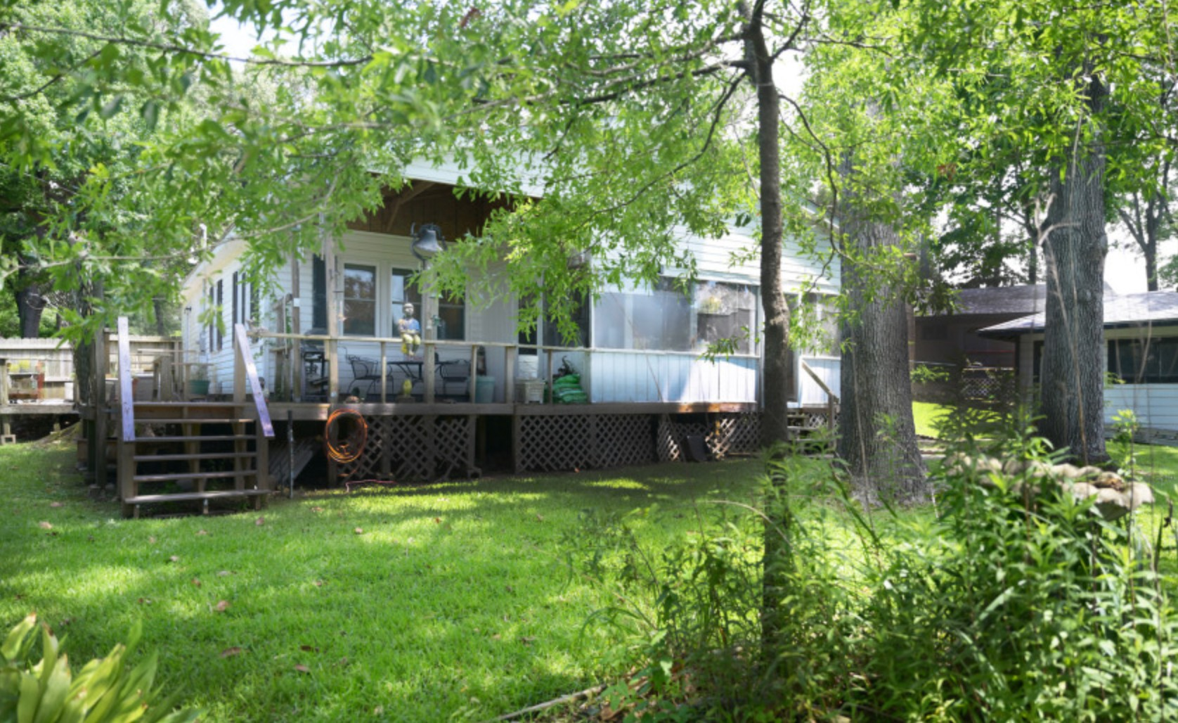Backyard view of a house with a wooden deck, surrounded by green trees and grass, with stairs leading up to the deck.