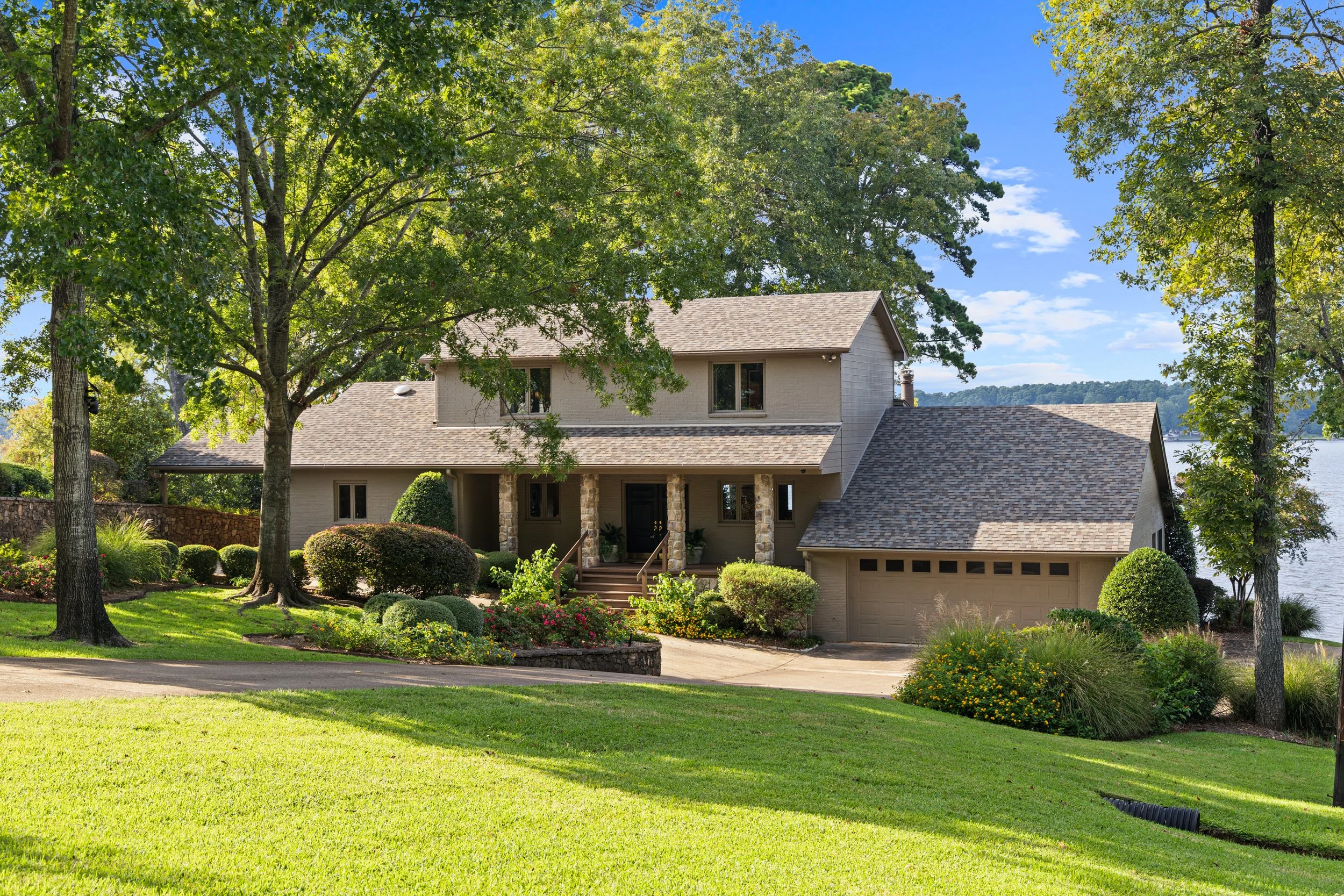 A large, two-story house with a front porch, surrounded by well-maintained lawn and landscaping, with multiple trees and a lake visible in the background.