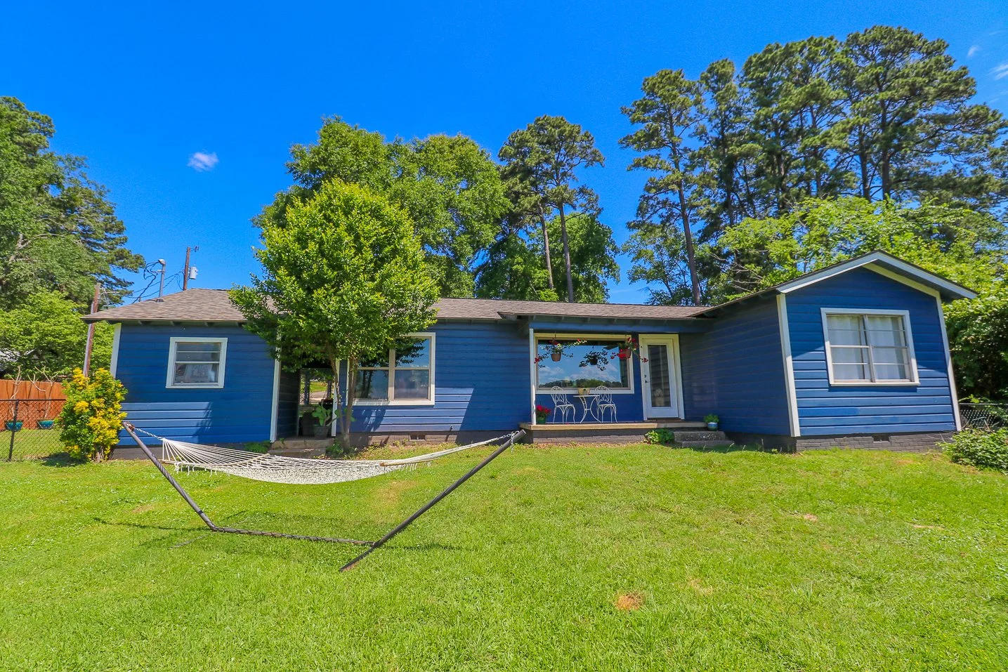 Blue house with a fenced backyard, hammock, and outdoor seating area, surrounded by trees on a sunny day.