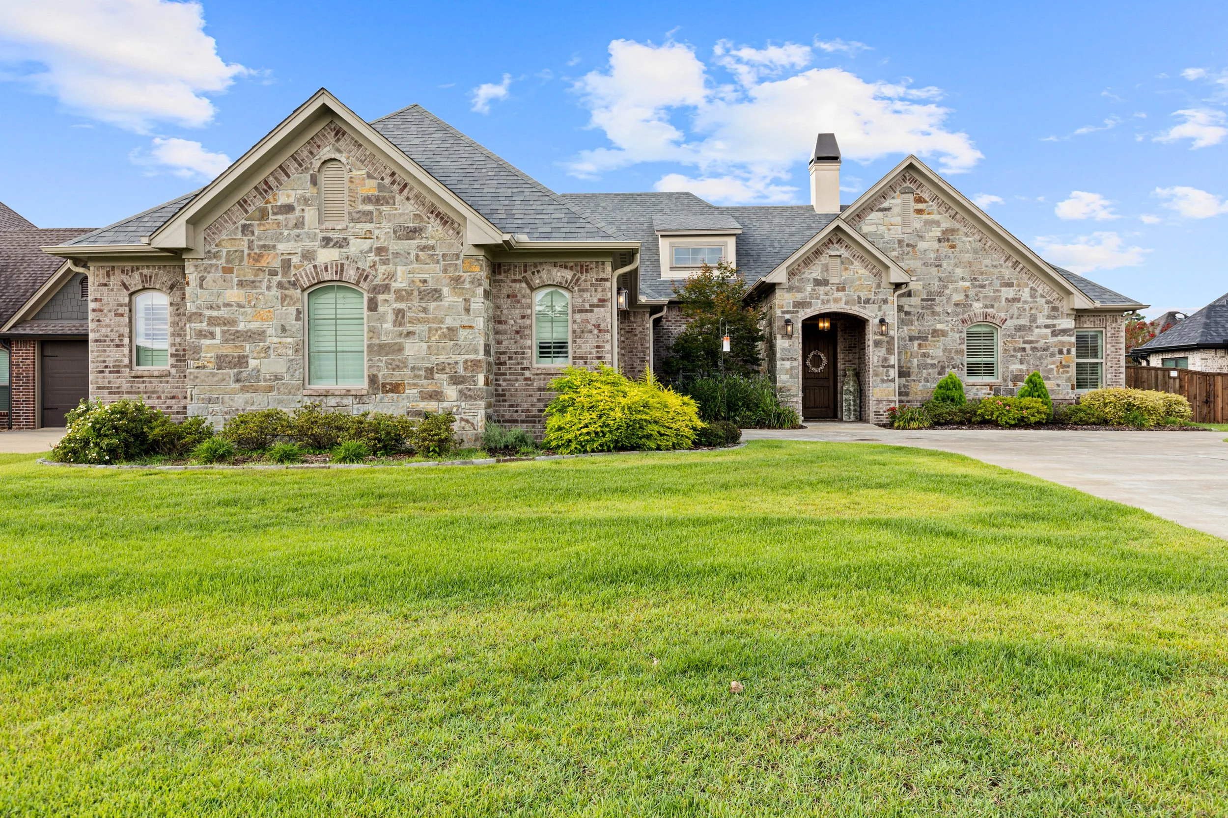 A large, elegant house with a stone exterior, front lawn, and driveway, under a partly cloudy sky.