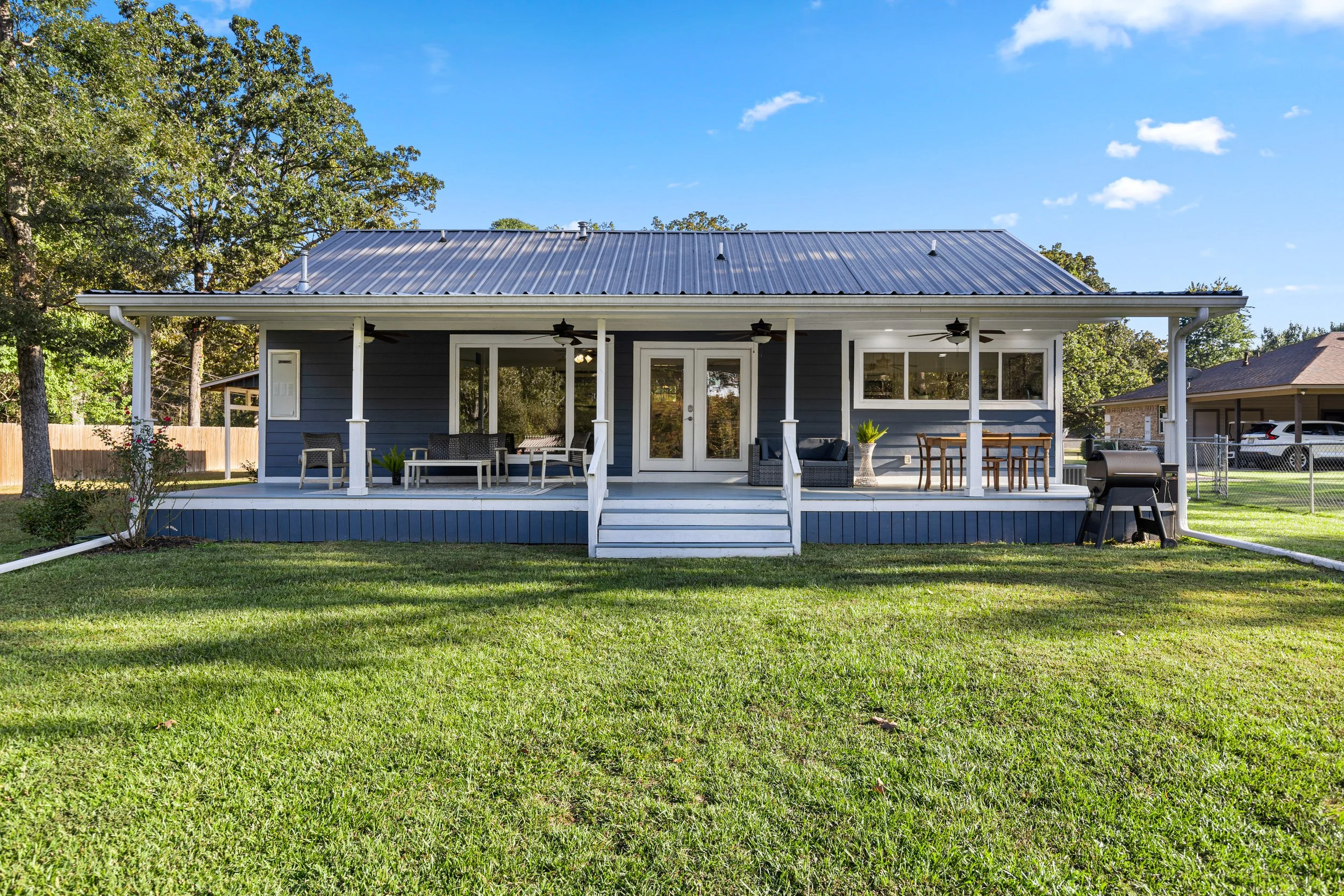 A blue house with a covered porch, white trim, and a metal roof, surrounded by a green lawn and trees in the background.