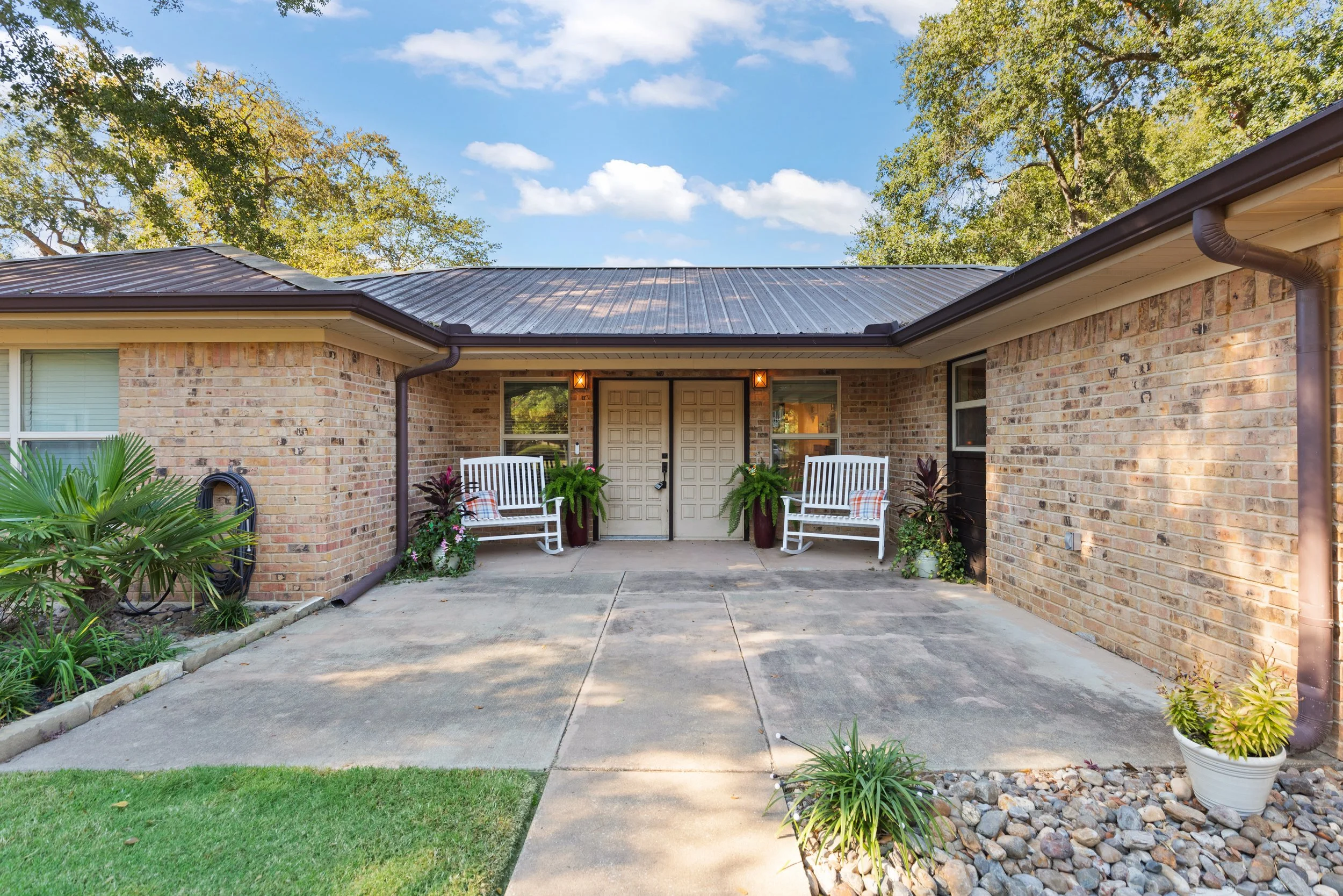 Front porch of a brick house with two white benches, potted plants, and a concrete walkway leading to the door under a metal roof with trees and a blue sky in the background.