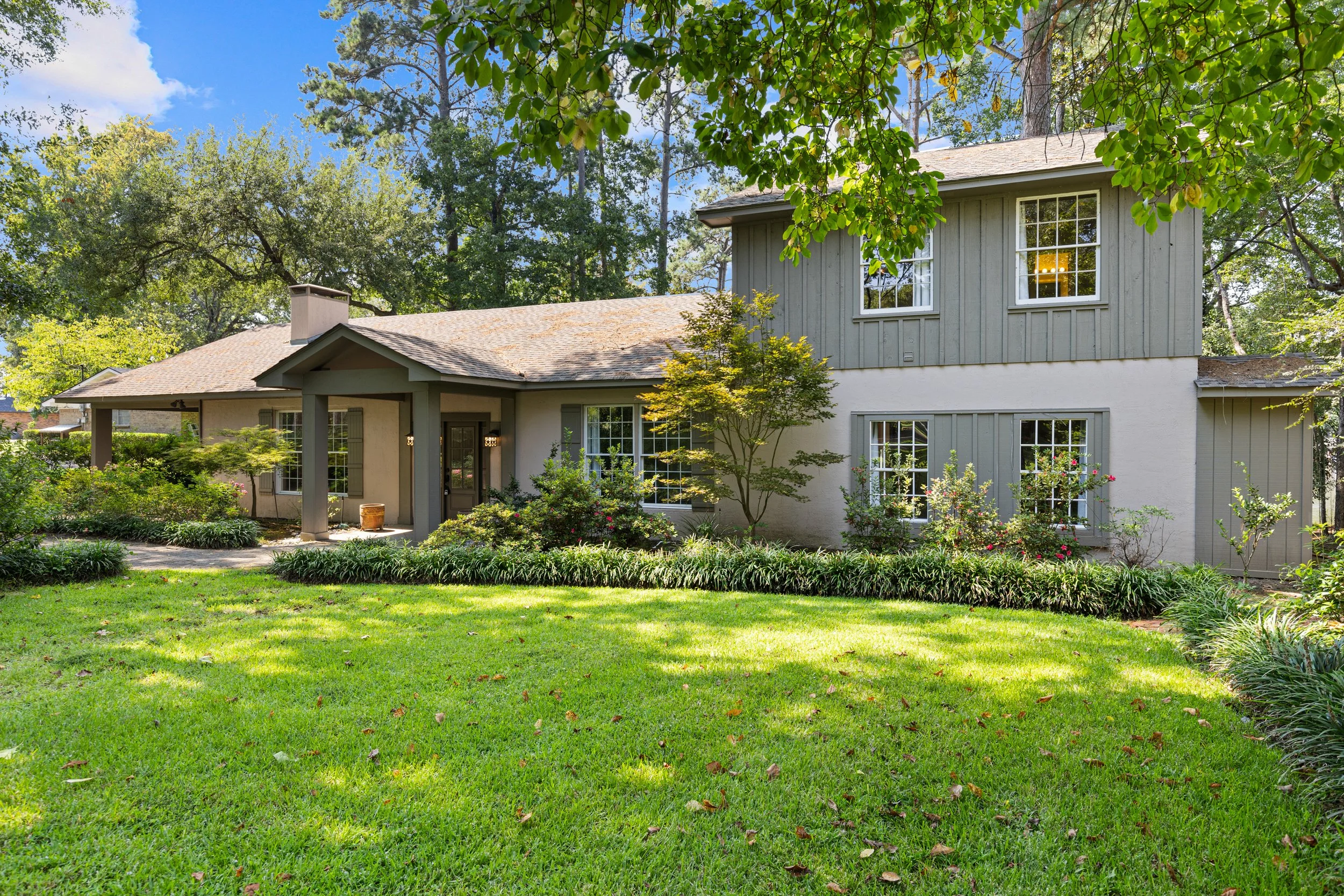 A two-story house with a front porch, surrounded by lush green lawn and trees, with windows and outdoor lighting, in a bright, sunny day.