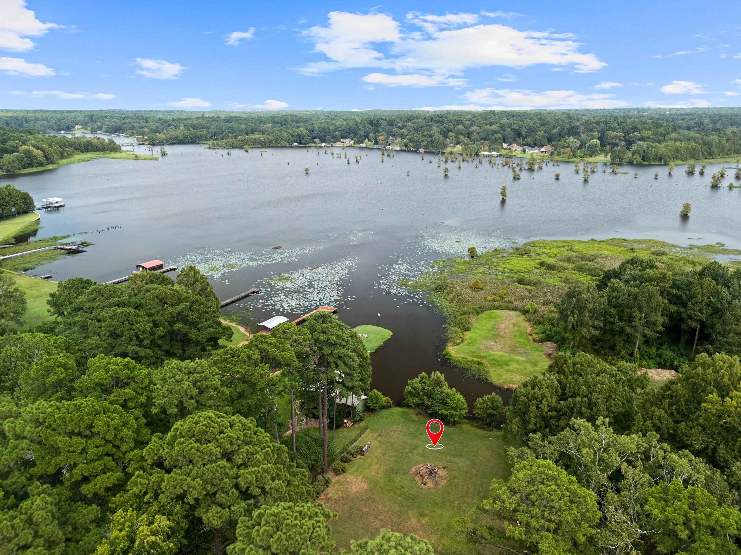 Aerial view of a large lake with surrounding green land, trees, and a few docks. The sky is partly cloudy.
