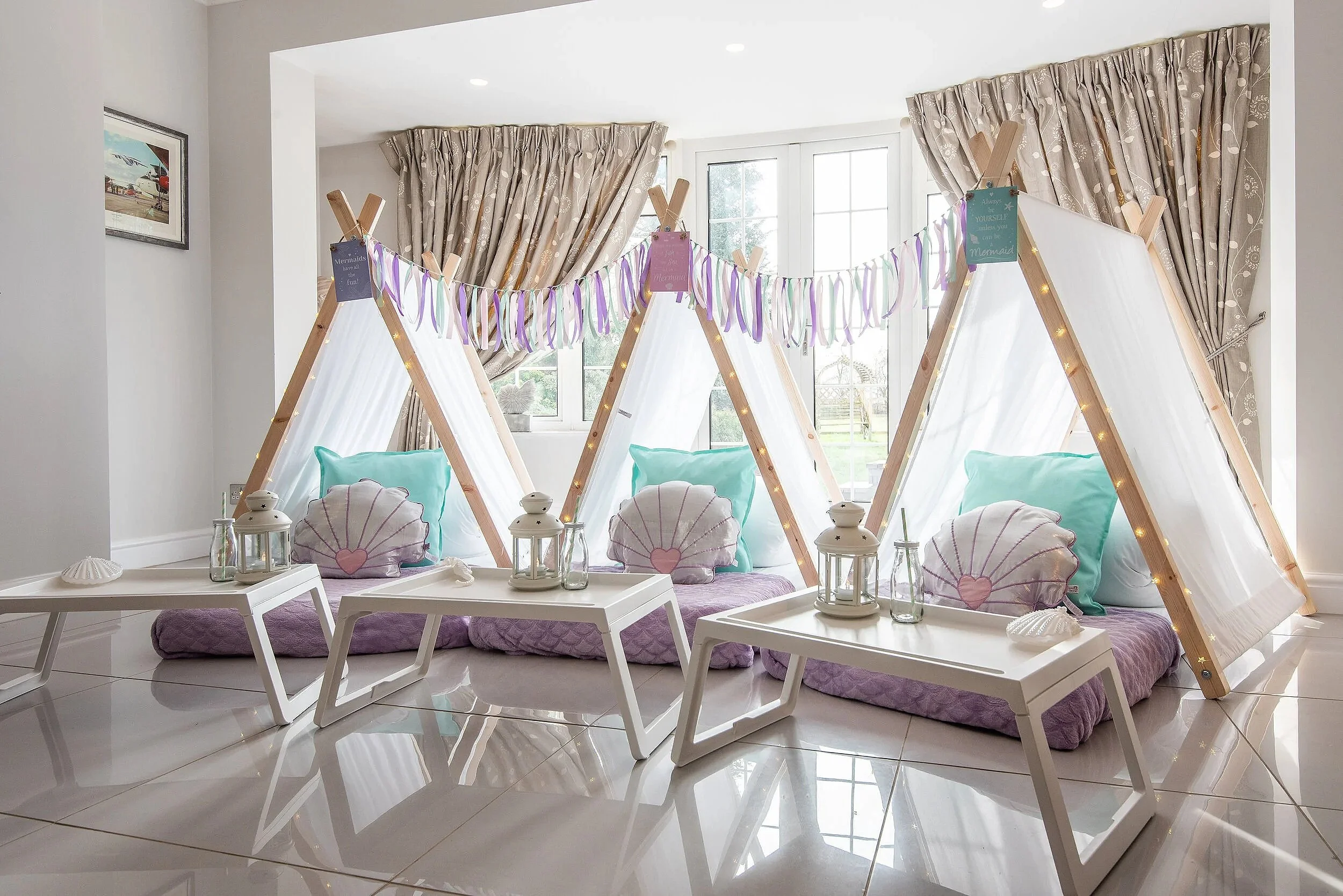 Indoor setting with three wooden teepees decorated with string lights and pastel-colored pillows, placed on a white tiled floor near large windows with beige patterned curtains, with white tables and lanterns in the foreground and a framed airplane picture on the wall.