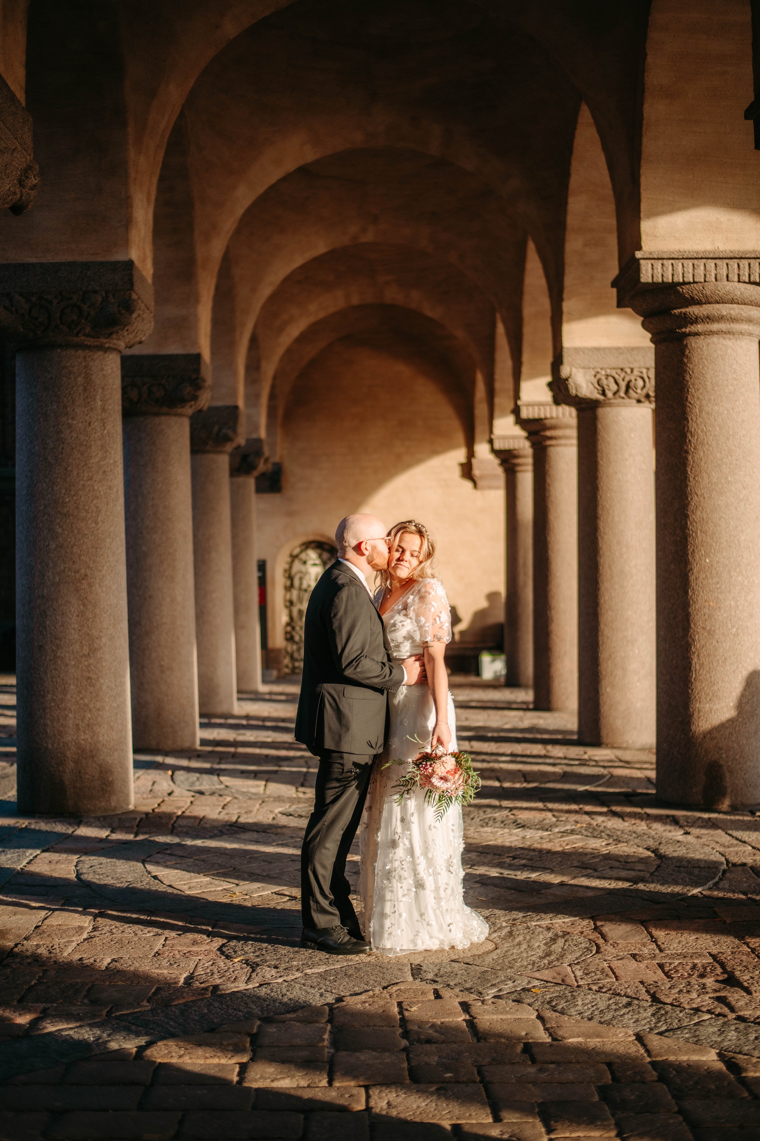 En brud och en brudgum i en tunnel med arkader, de står nära varandra och delar ett ögonblick, bruden håller en bukett blommor, kvällen är varm och ljuset är gyllene.