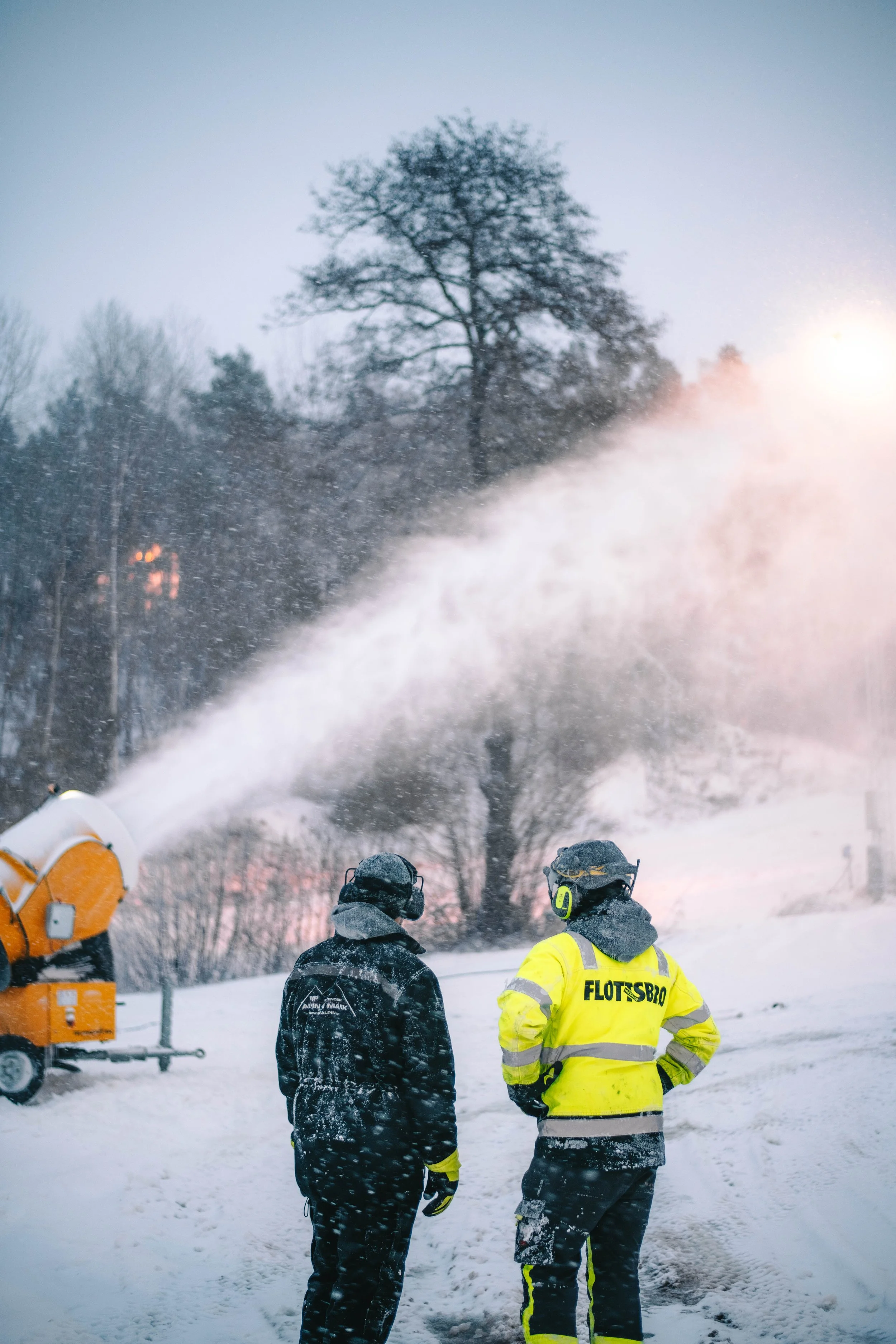 Två personer i vinterkläder, en i svart och en i gul, övervakar en snöskjutare som sprutar snö. Bakom står ett träd och det är snö på marken.