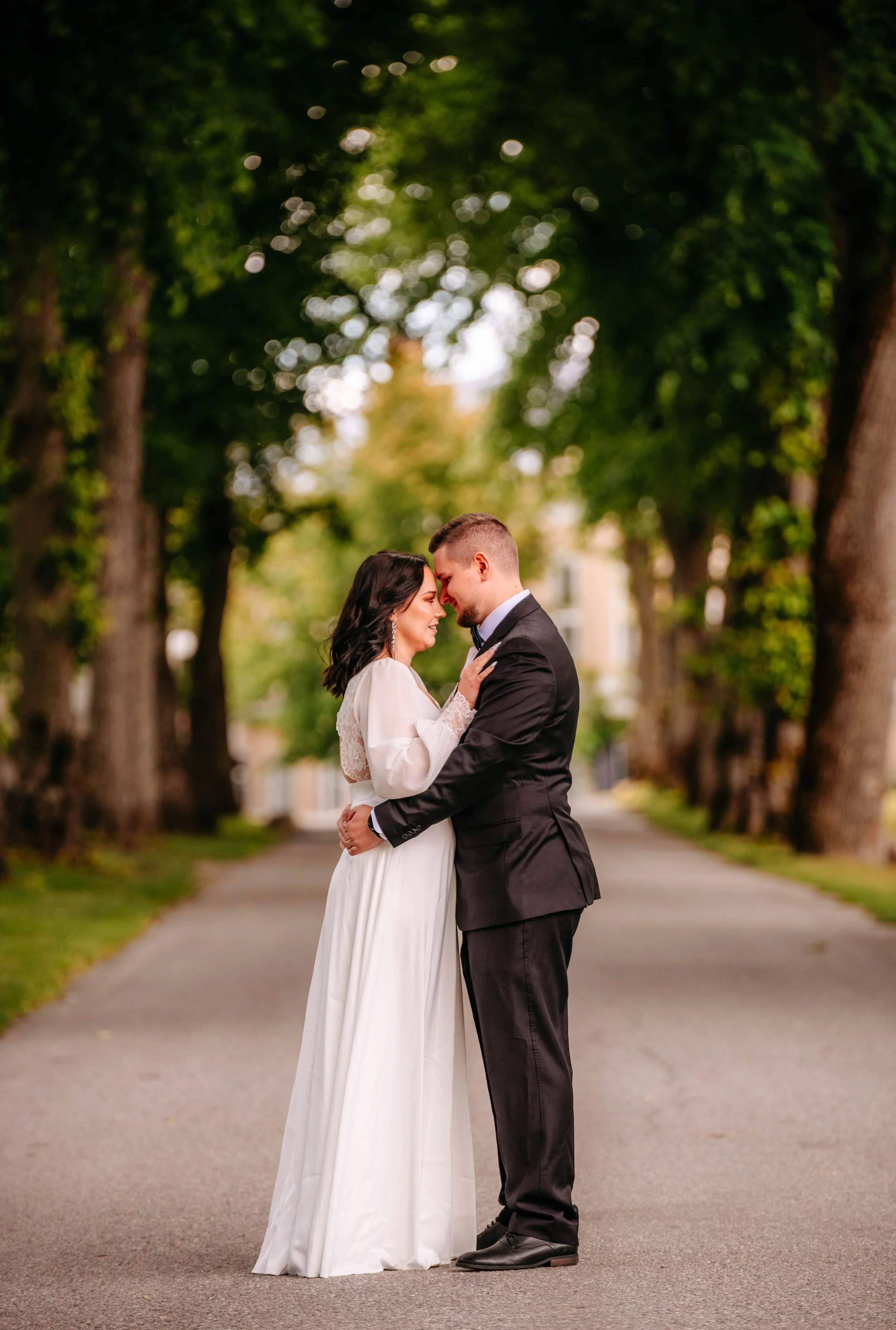 En brud och en brudgane står nära varandra på en gata omgiven av träd, svarta kläder. De lutar sina huvuden mot varandra och ler, i en romantisk pose i skogsmiljö.