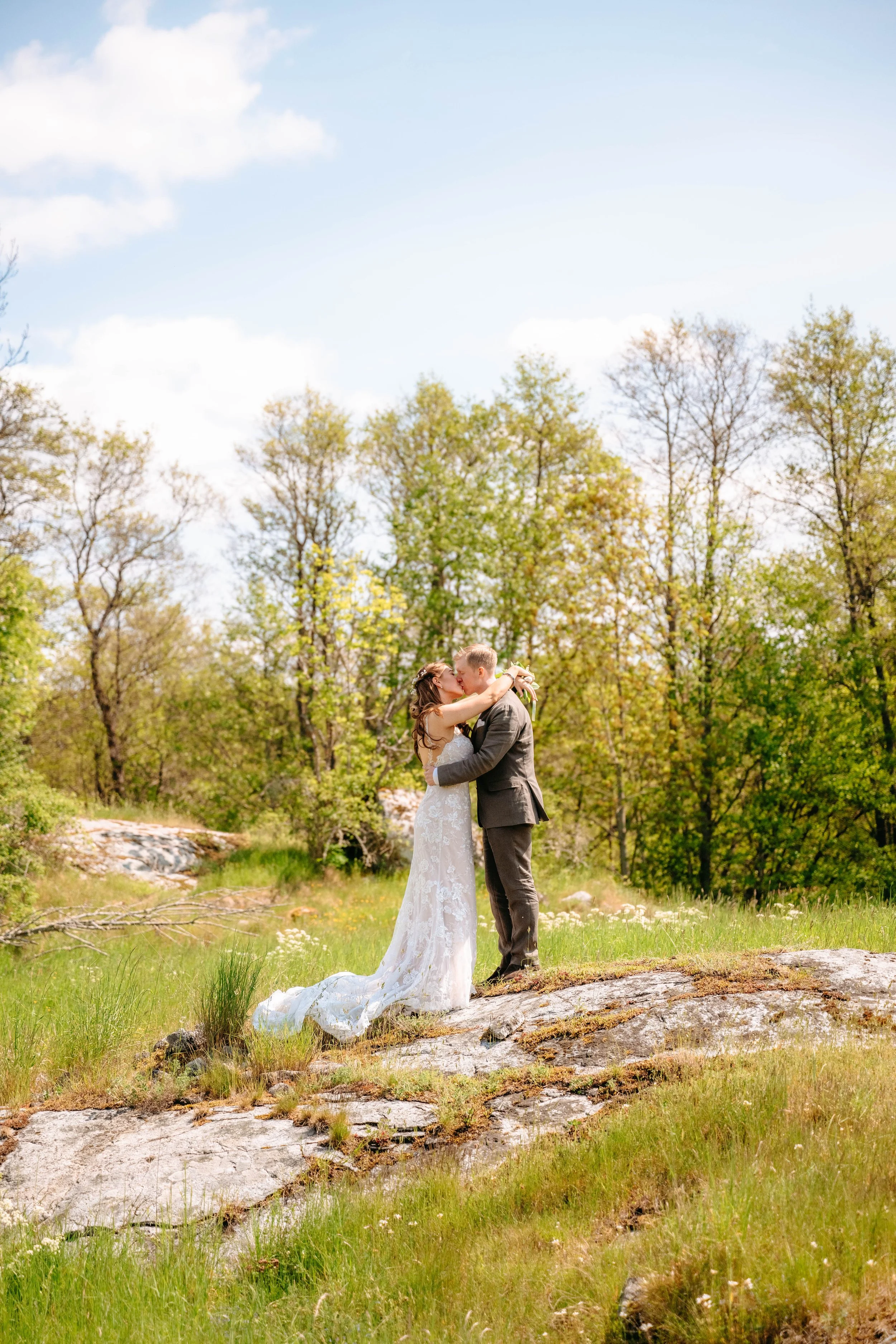 En brud och brudg sonar i en skogig äng, omfamnar och kysser varandra, under en klar himmel.