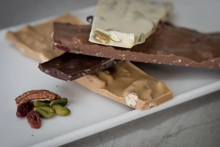 Assorted types of chocolate bars on a white platter, with some pistach Nuts and dried cranberries on the side.