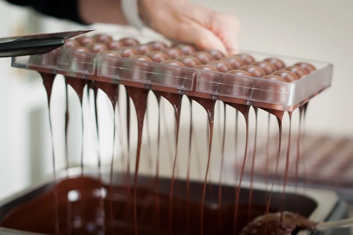 Hand pouring melted chocolate over a tray of round chocolate candies.