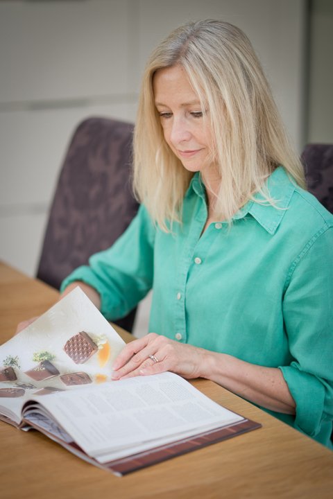 A woman with shoulder-length blonde hair wearing a green button-up shirt sitting at a table, looking at an open magazine with images of chocolate and desserts.
