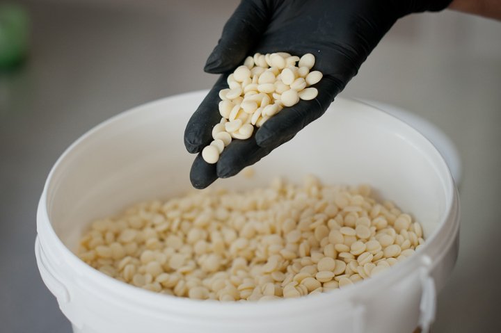A person wearing a black glove holding white chocolate chips above a white bucket filled with more white chocolate chips.