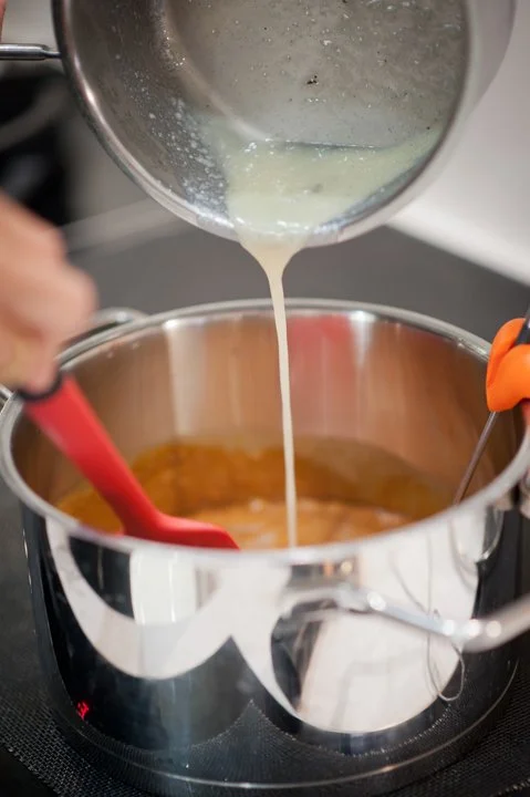 Pouring a creamy liquid into a saucepan on a stove, with a red spatula and orange tongs nearby.