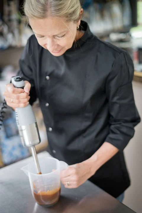 A woman in a black chef's coat blending a liquid in a clear measuring cup using an immersion blender in a kitchen setting.