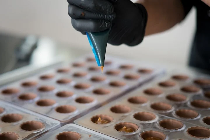 A person wearing black gloves piping chocolate into mold cavities on a silicone baking mat for making chocolates.