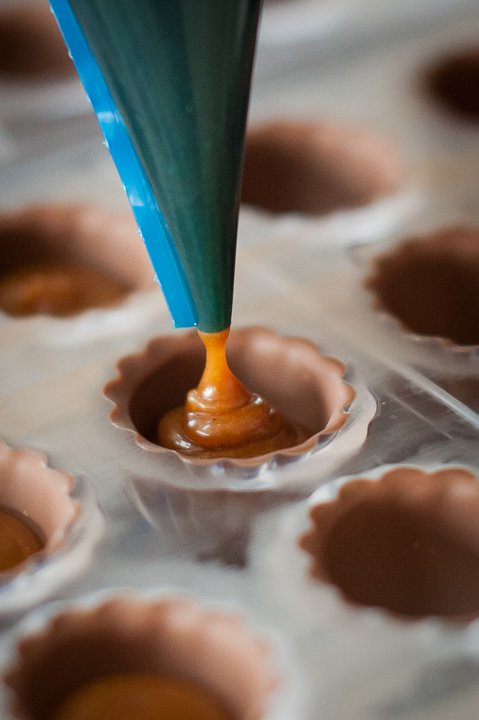 A piping bag filling chocolate into mini tart shells on a baking sheet.