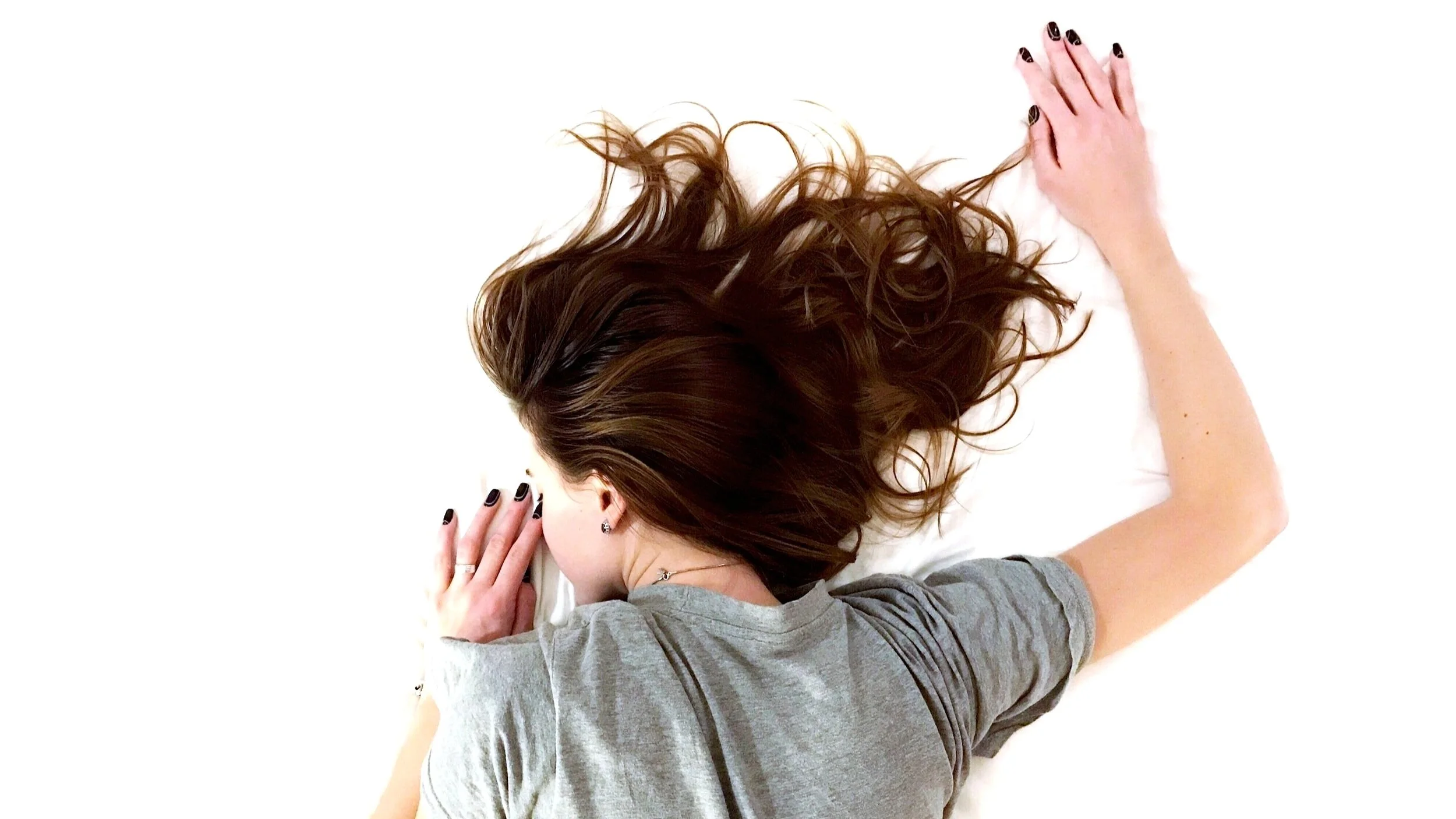 A woman with long brown hair is lying down on her side, resting her head on a white surface, wearing a gray t-shirt, with her hand near her face and her other arm extended outward.