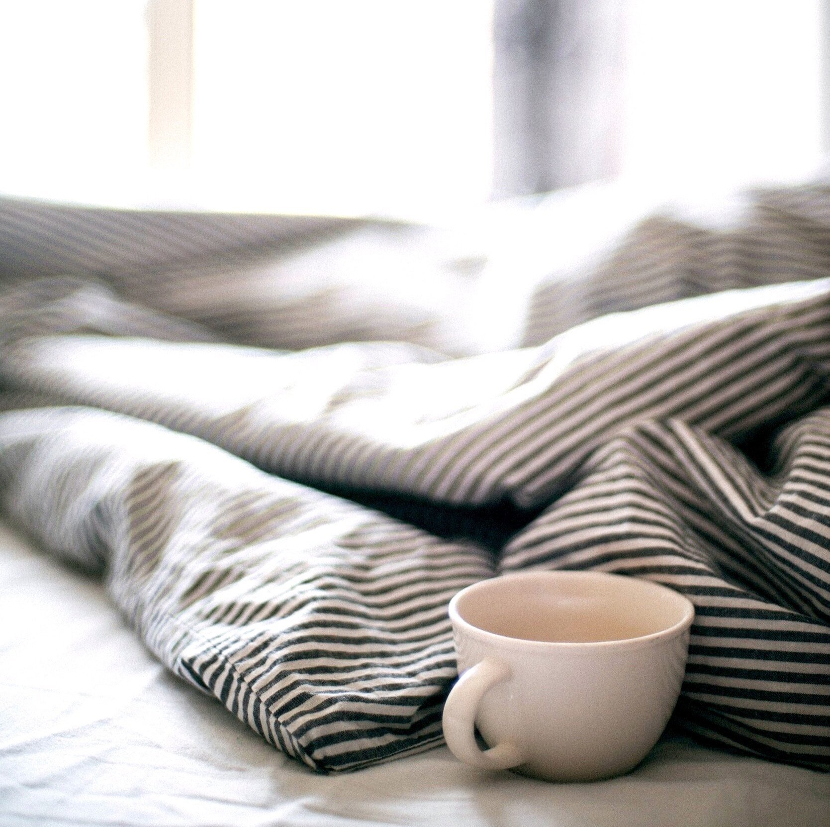 A white mug with a handle resting on a white bed nearby a loosely placed black and white striped bedsheet, with soft natural light coming through a window in the background.