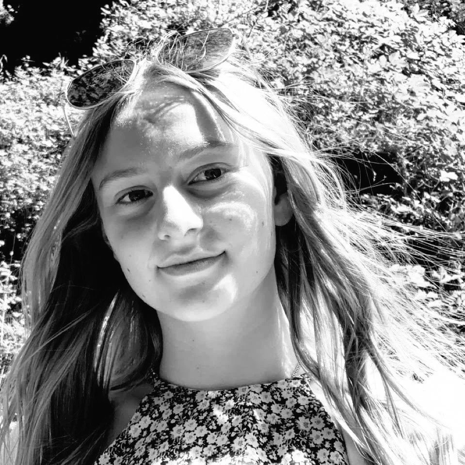 Black-and-white photo of a young woman outdoors with sun on her face, wearing sunglasses on her head and a floral top.