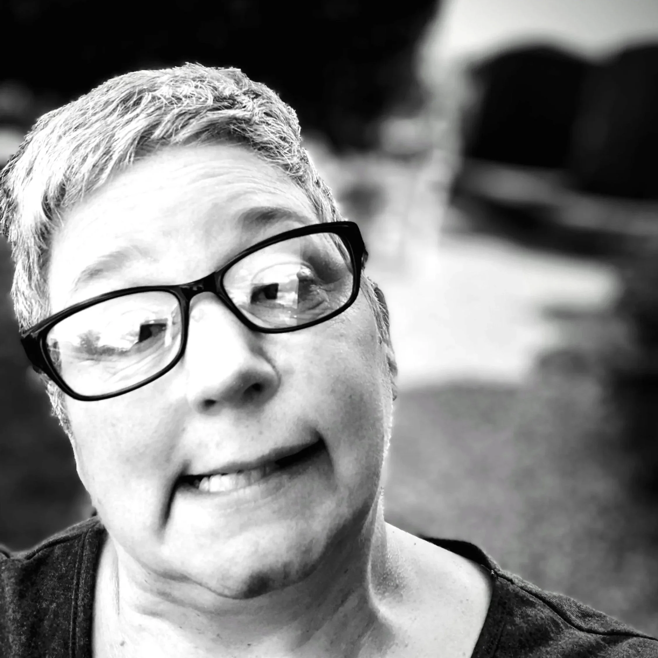 Black and white close-up photo of a person with short hair, wearing glasses, and smiling outdoors.