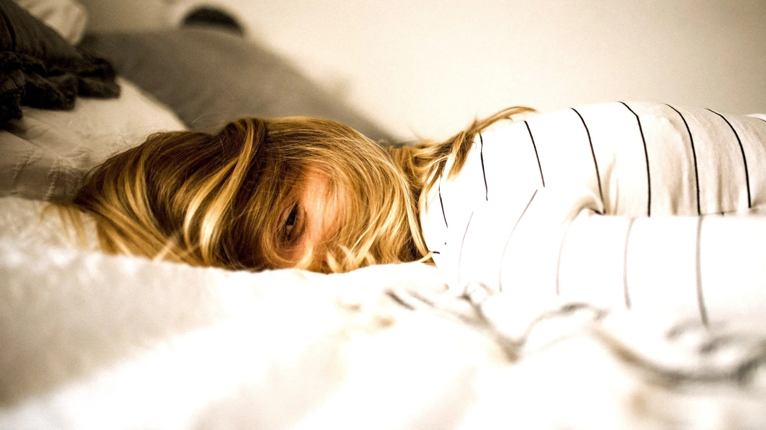 A person with blonde hair lying face down on a bed, wearing a white shirt with black horizontal stripes.